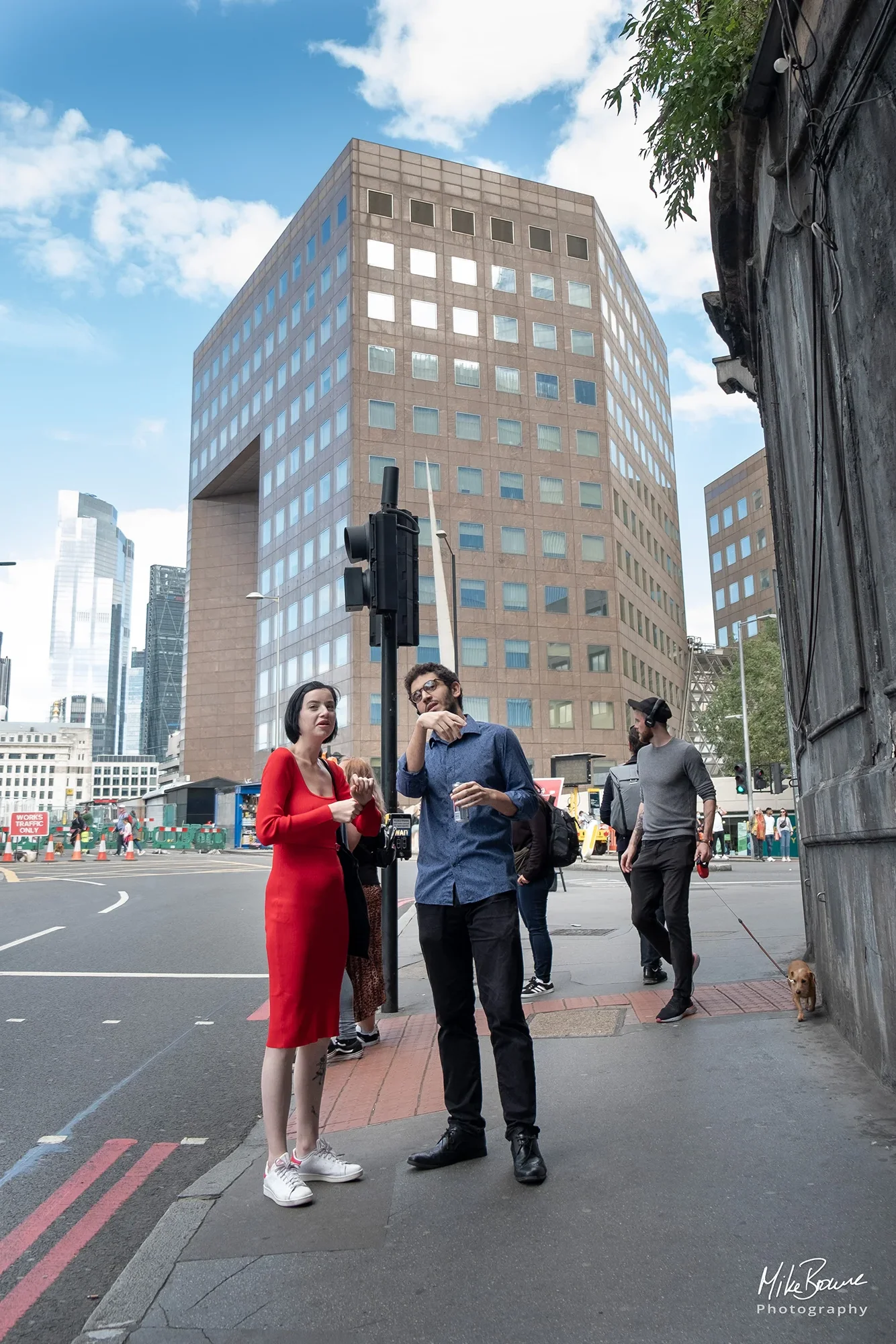 Woman in a red dress and white trainers conversing with a man stood by traffic lights in front of modernist building