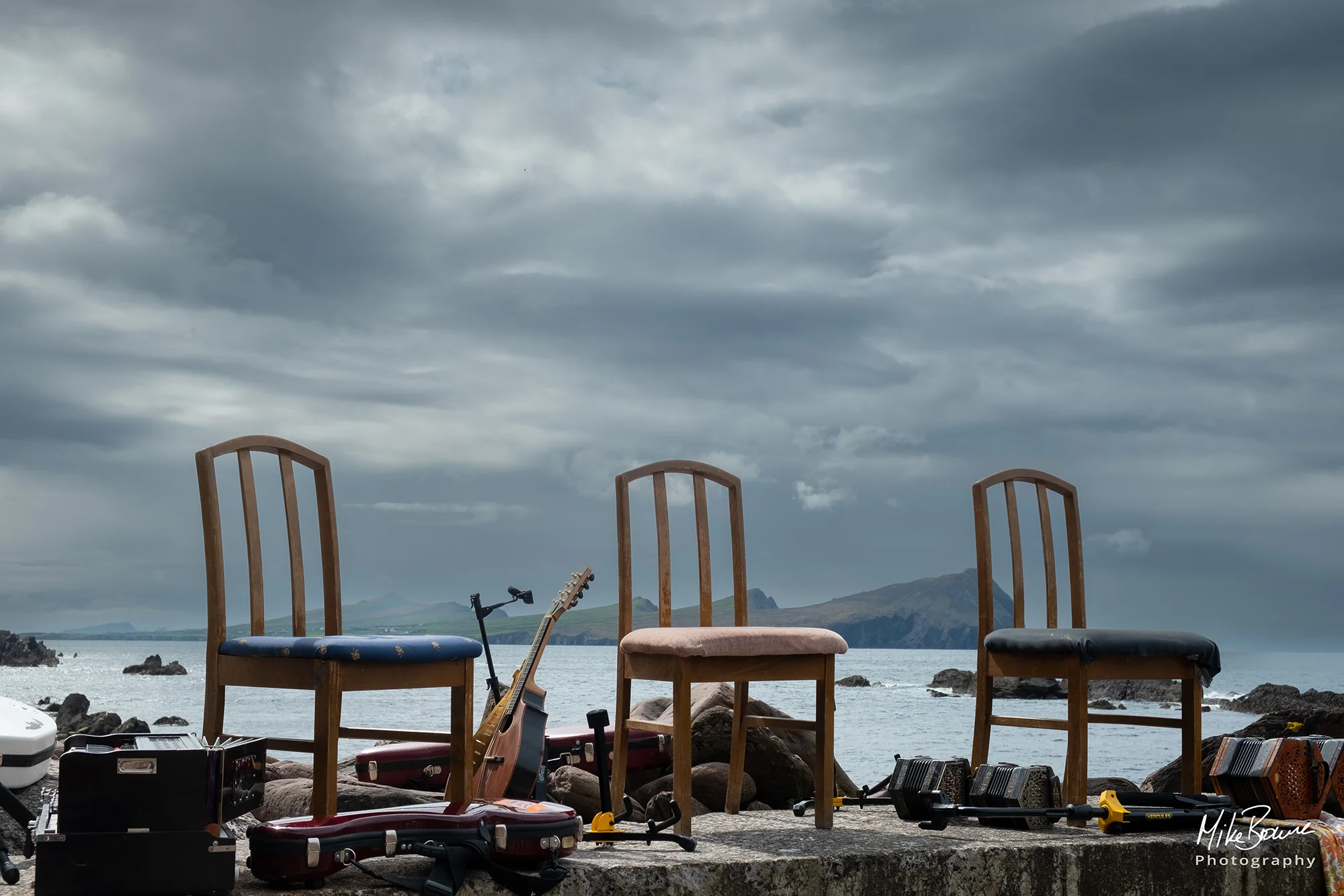 Three empty chairs and musical instruments on harbour wall with mountains beyond