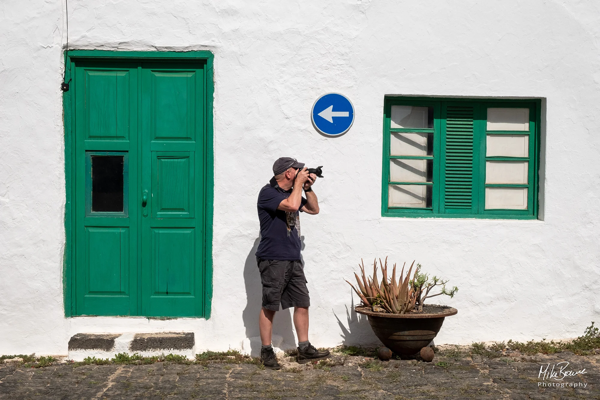 Man with camera taking a photo in front of a white wall with a blue arrow pointing in opposite direction in Teguise, Lanzarote