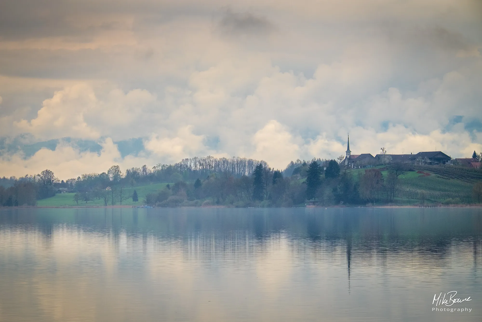 Hazy clouds over lake with mist on shoreline blurring church spire and trees