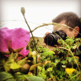 Behind a pink flower and foliage a worksop attendee focuses on a shot