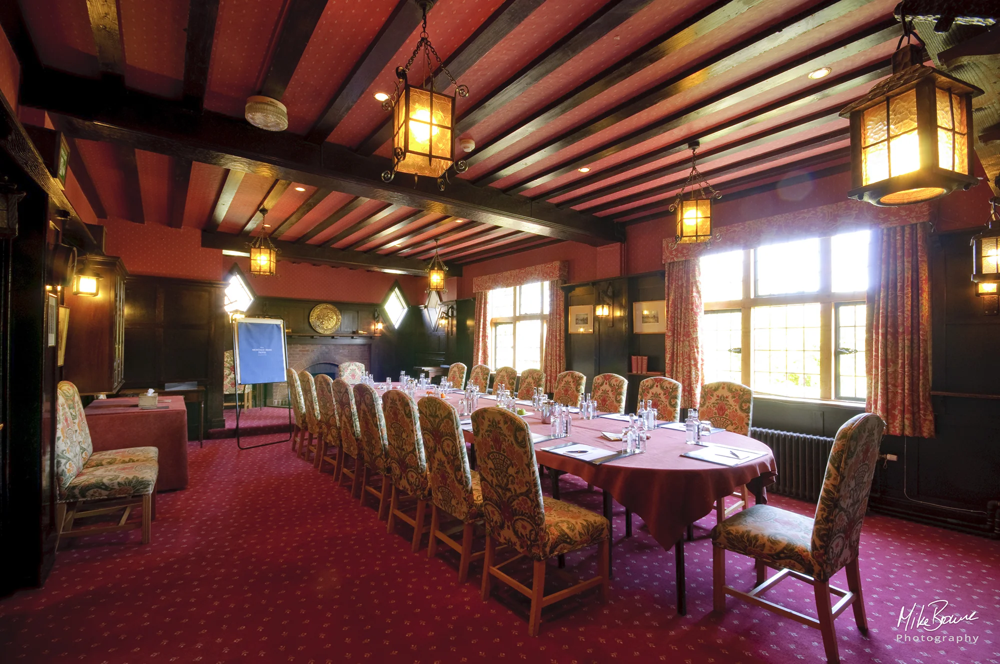 Wood panelled function room with bright windows set up with long table, flip chart and chairs for a business meeting of 18 people