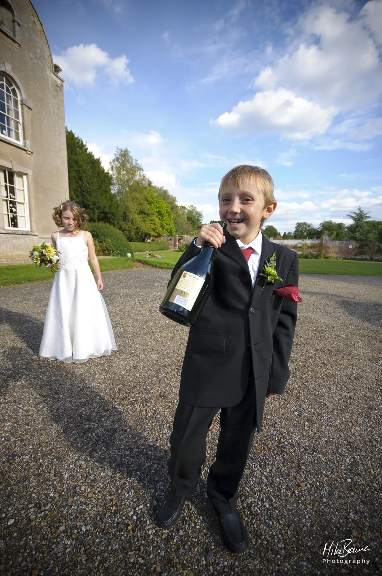 Young grinning page boy with a bottle of Champagne outside wedding reception being watched by a Bridesmaid