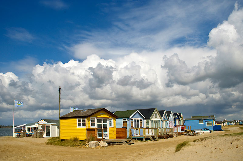 Bright yellow beach hut with huts behind and a sky of cumulus clouds