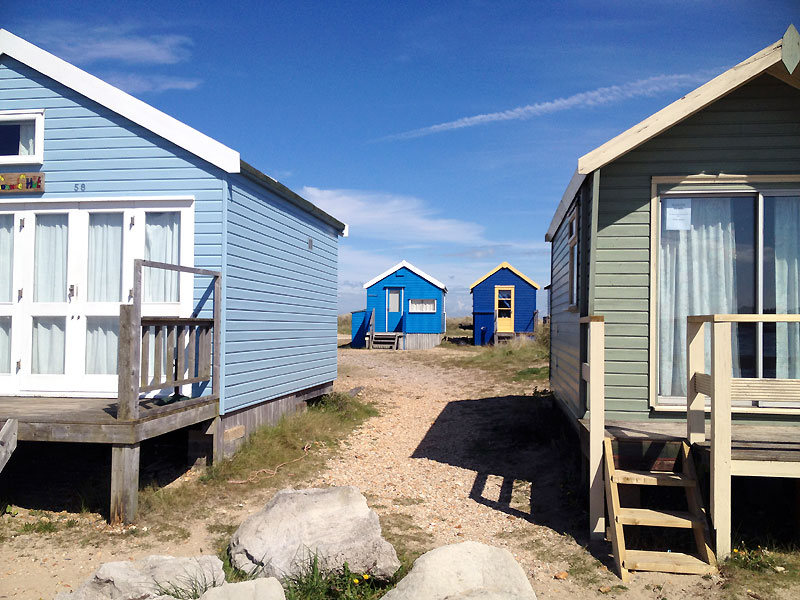 Four beach huts with two in the background and two in the four ground and a sandy path between with blue sky and cirrus clouds
