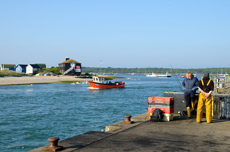 Fisherman standing to the right on the dock whilst Mudeford ferry crosses the water