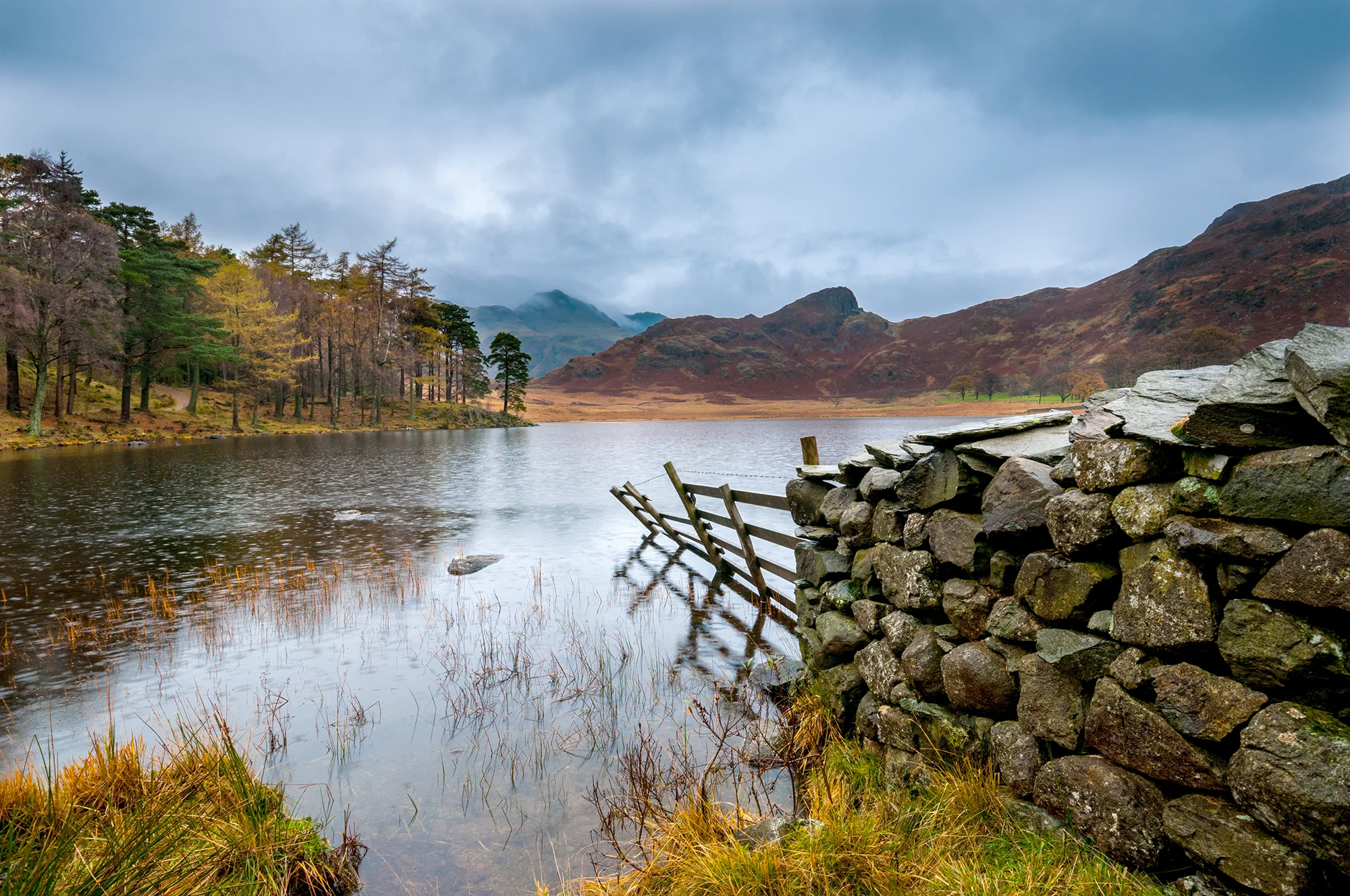 Stone wall leading into a lake with forest and cloud topped hills in distance on a rainy day