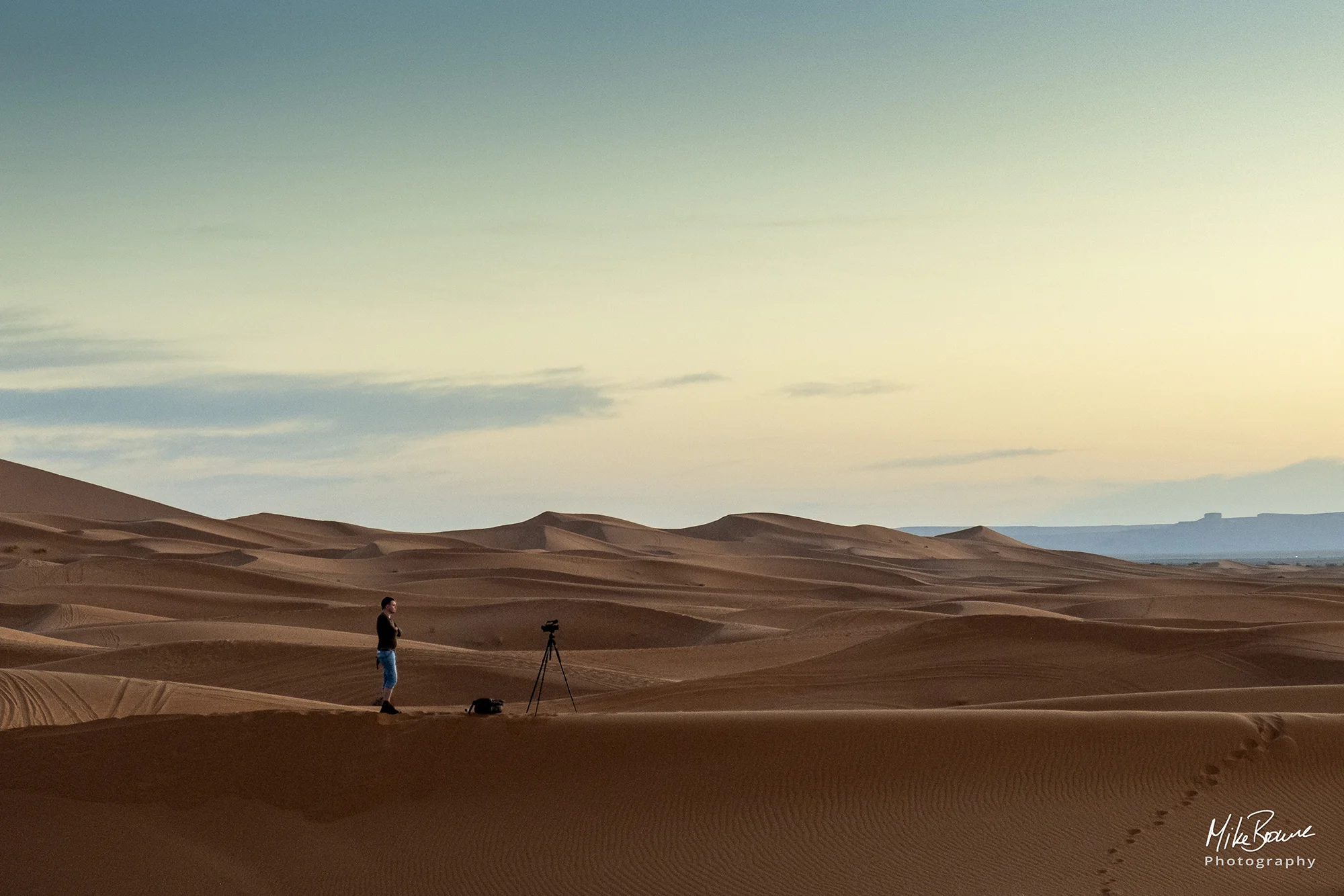 Man with camera on tripod standing amongst sand dunes in a desert at sunset