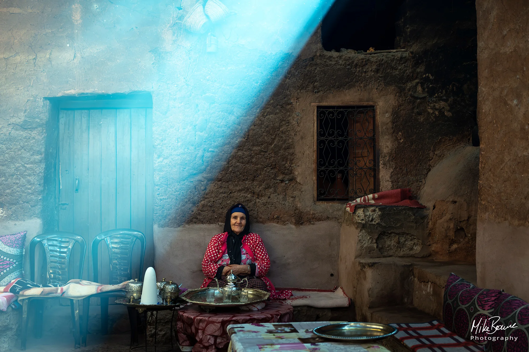 Elderly woman seated in a dim, smoke-filled rough walled room, illuminated by a beam of light.