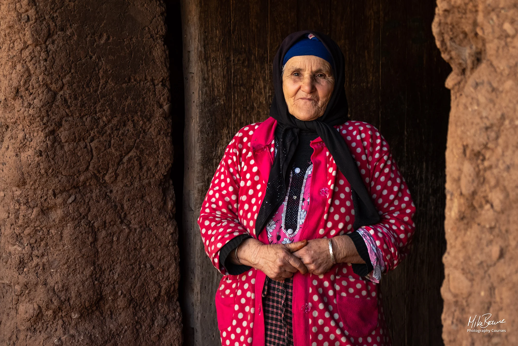 Elderly woman wearing red dress and hijab standing in doorway in village in Atlas Mountains of Morocco