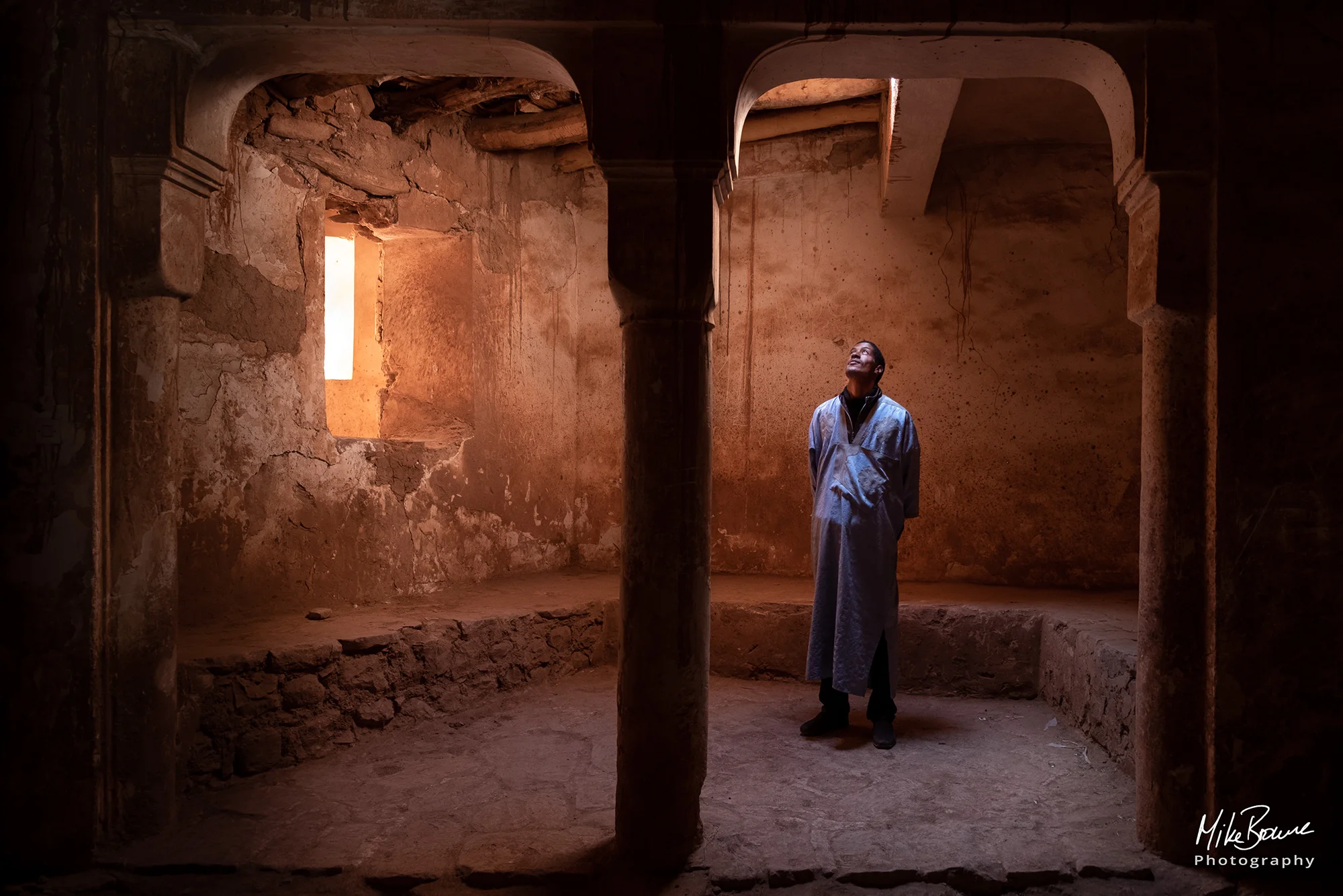 Man standing between pillars and looking up at overhead window in a dimly lit Kasbah in Morocco