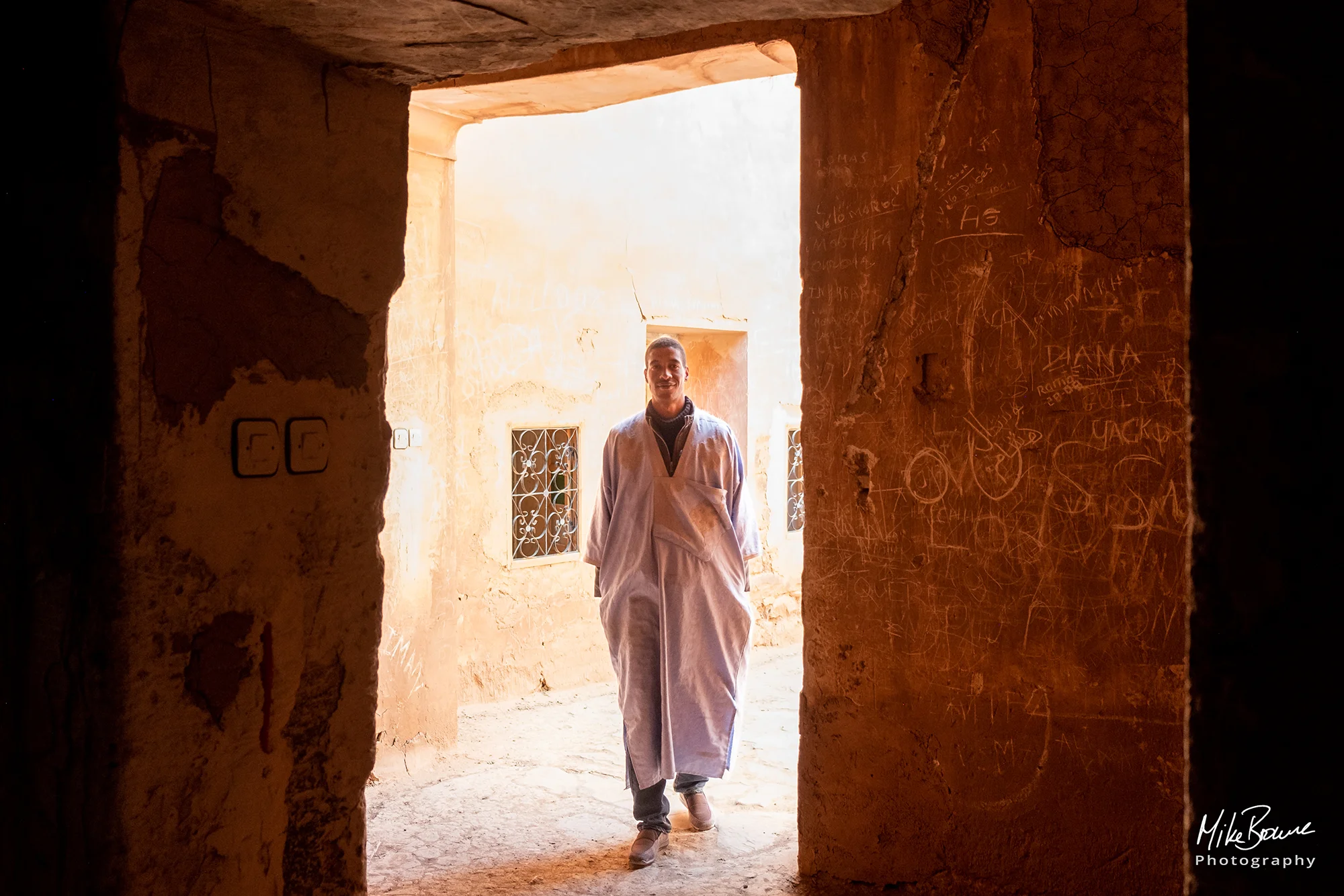 Young man wearing a grey djellaba walking in from the sunshine through a doorway in a Kasbah in Morocco