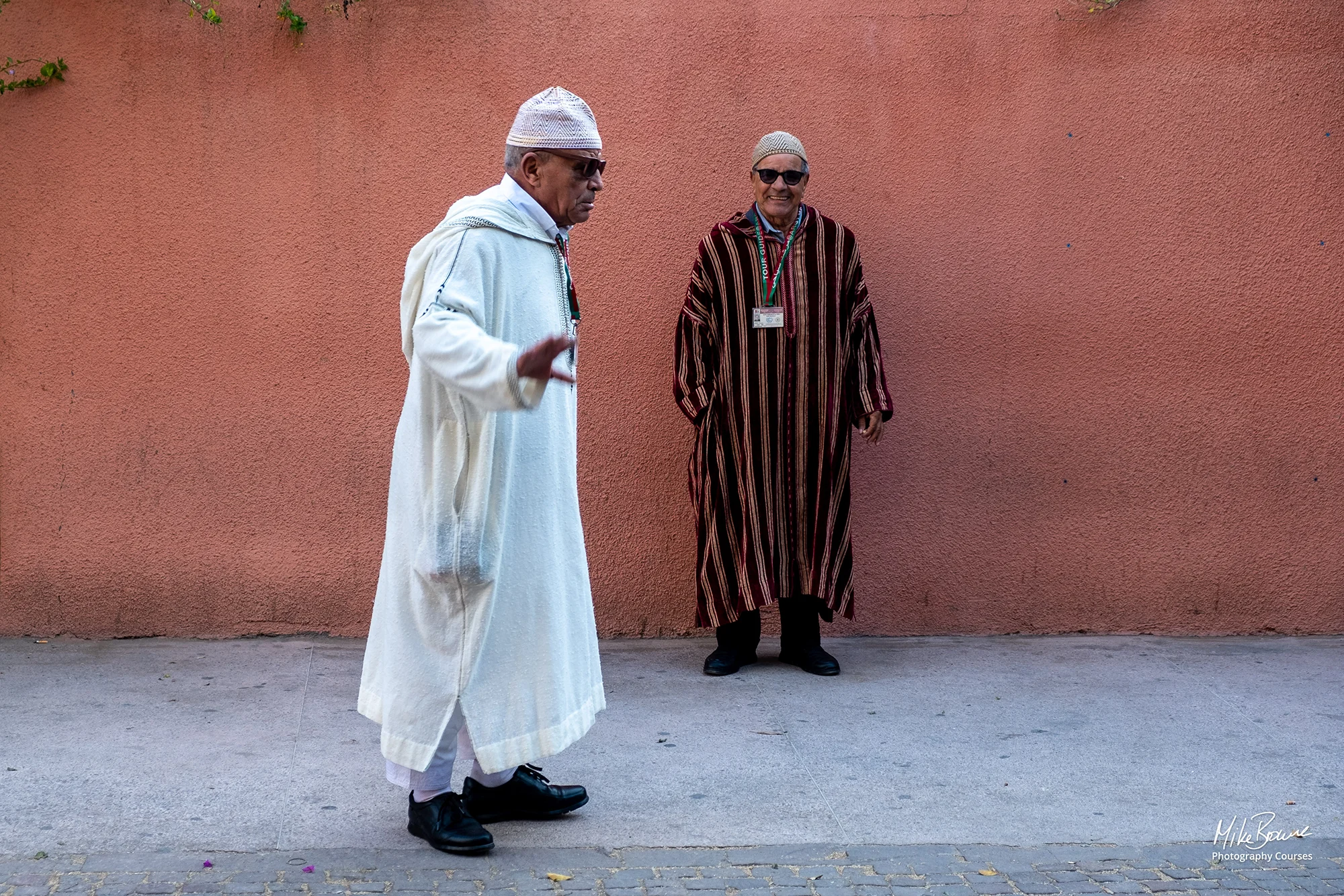 Man wearing white Moroccan djellaba and sunglasses gesturing as he walks past. Friend behind is smiling