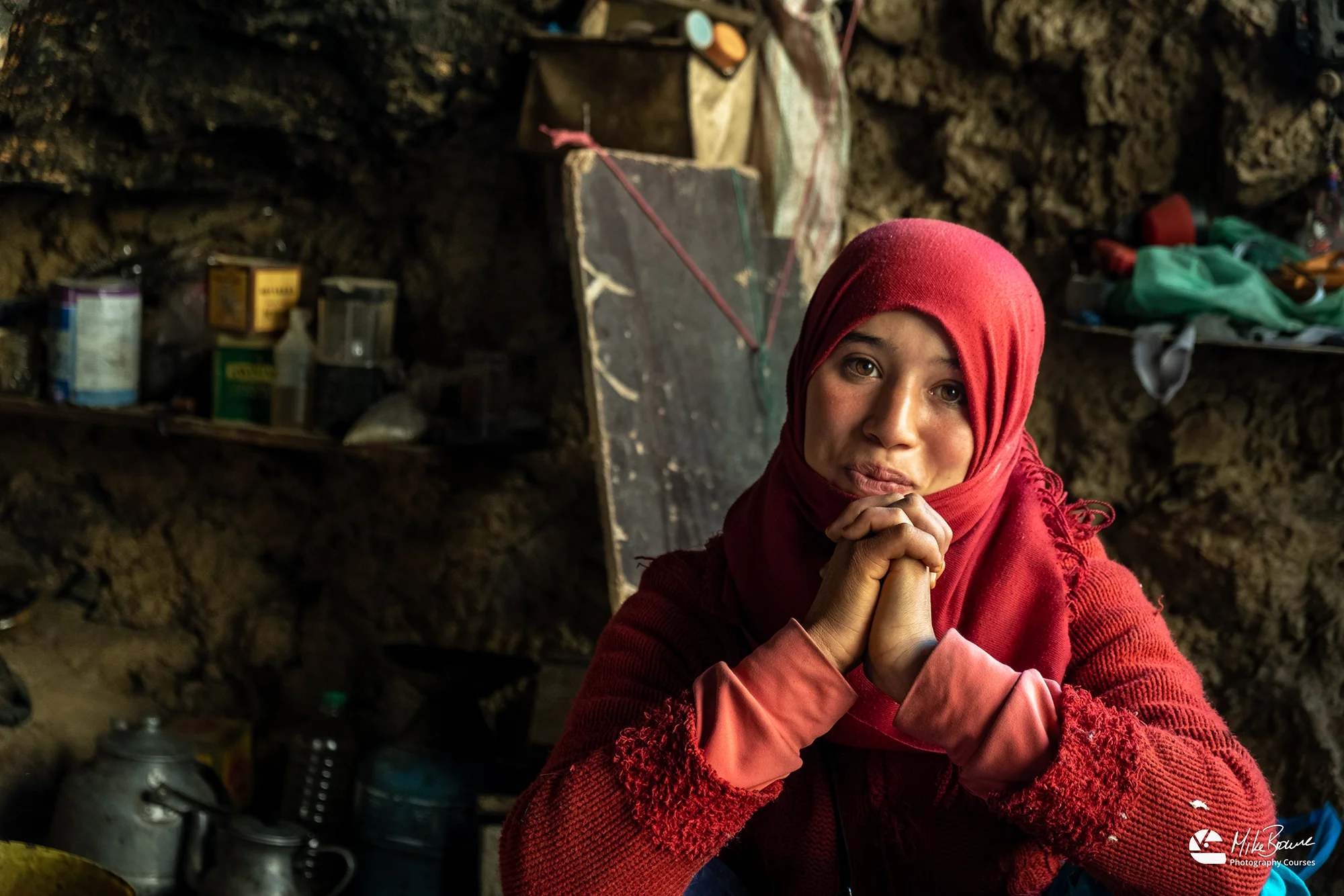 Young smiling Muslim girl wearing red hijab with shelves and tea kettle on leaning shelves behind her in a cave in the Todra Gorge, Morocco