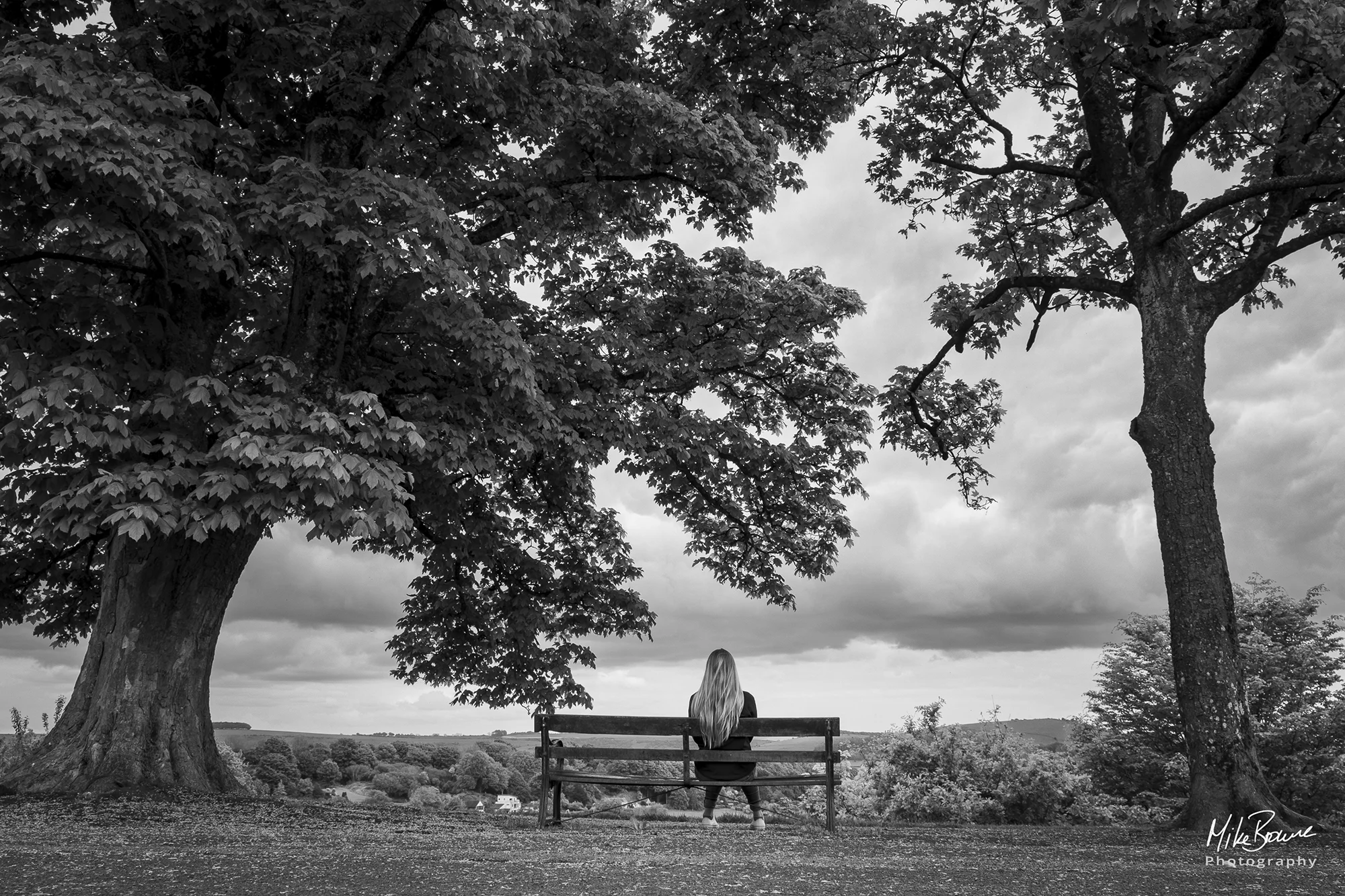 Woman sitting alone on a bench between two trees admiring cloudy sky over a valley