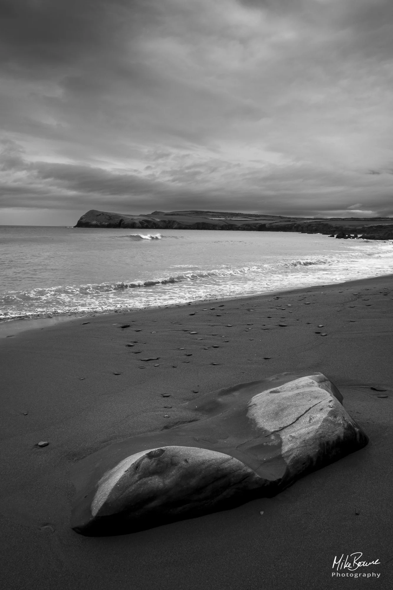 Curvy rock on a sand beach by the sea with splashing wave and headland beyond on a cloudy day