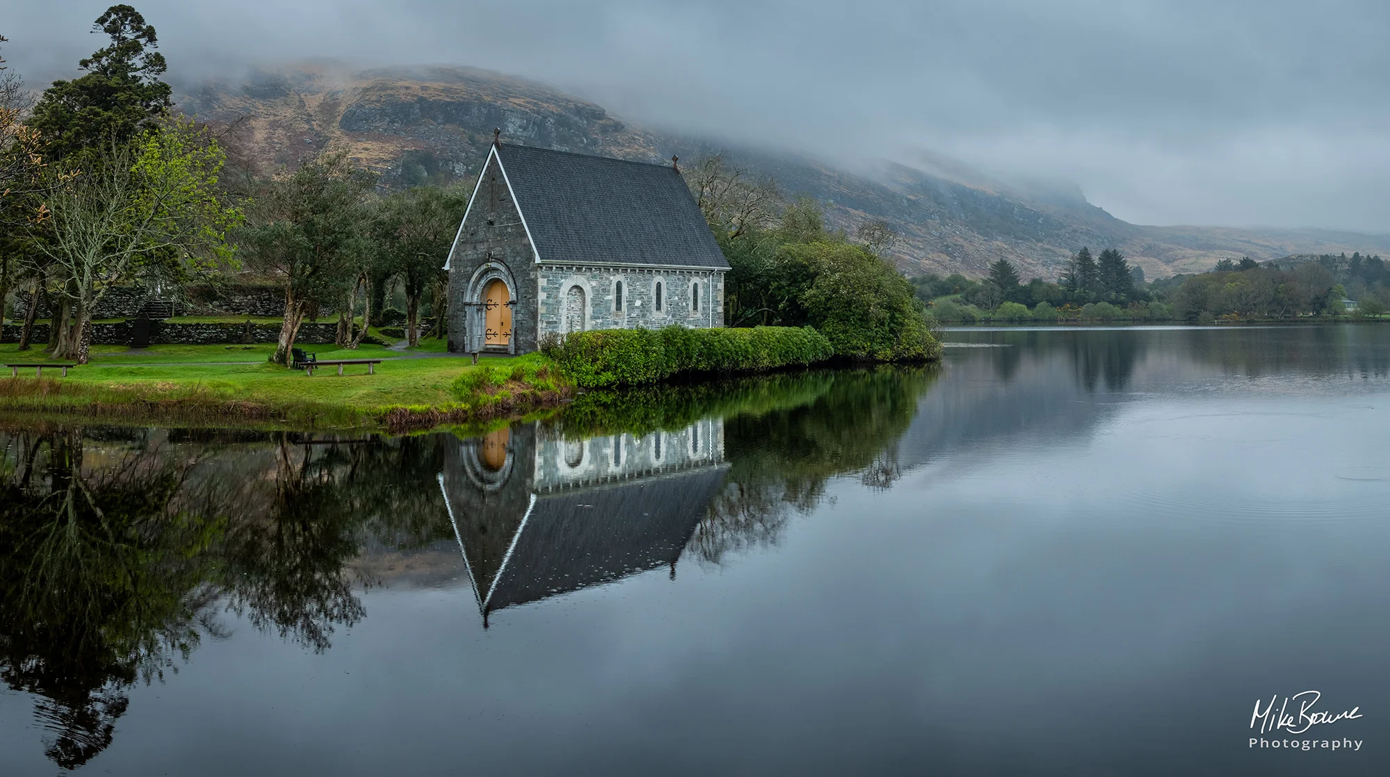 St Finbarr church surrounded by trees and misty mountain behind reflected in lake at Gougane Barra in Ireland