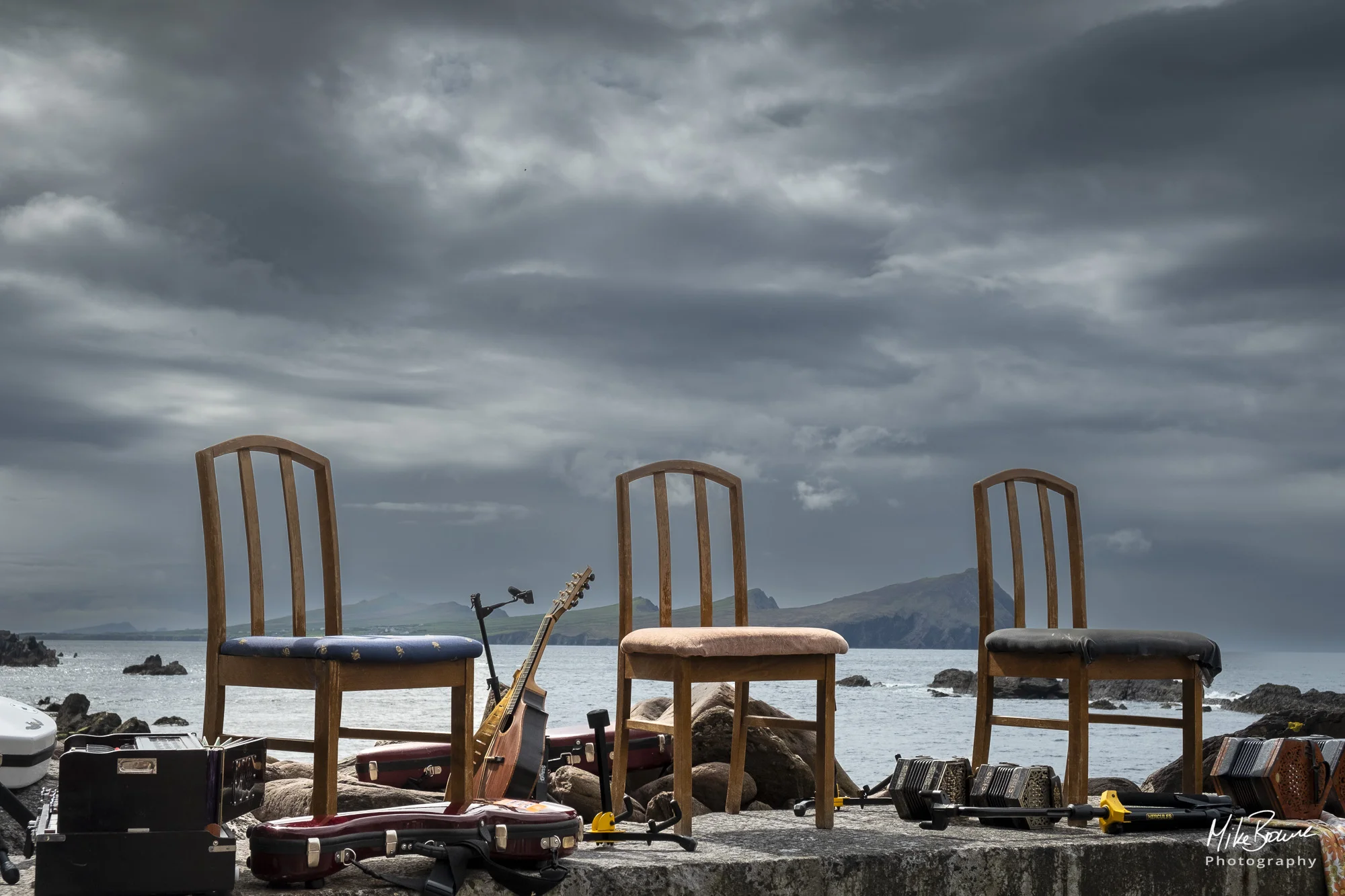 three chairs on a sea wall surrounded by musical instruments with storm clouds over sea and island in the distance