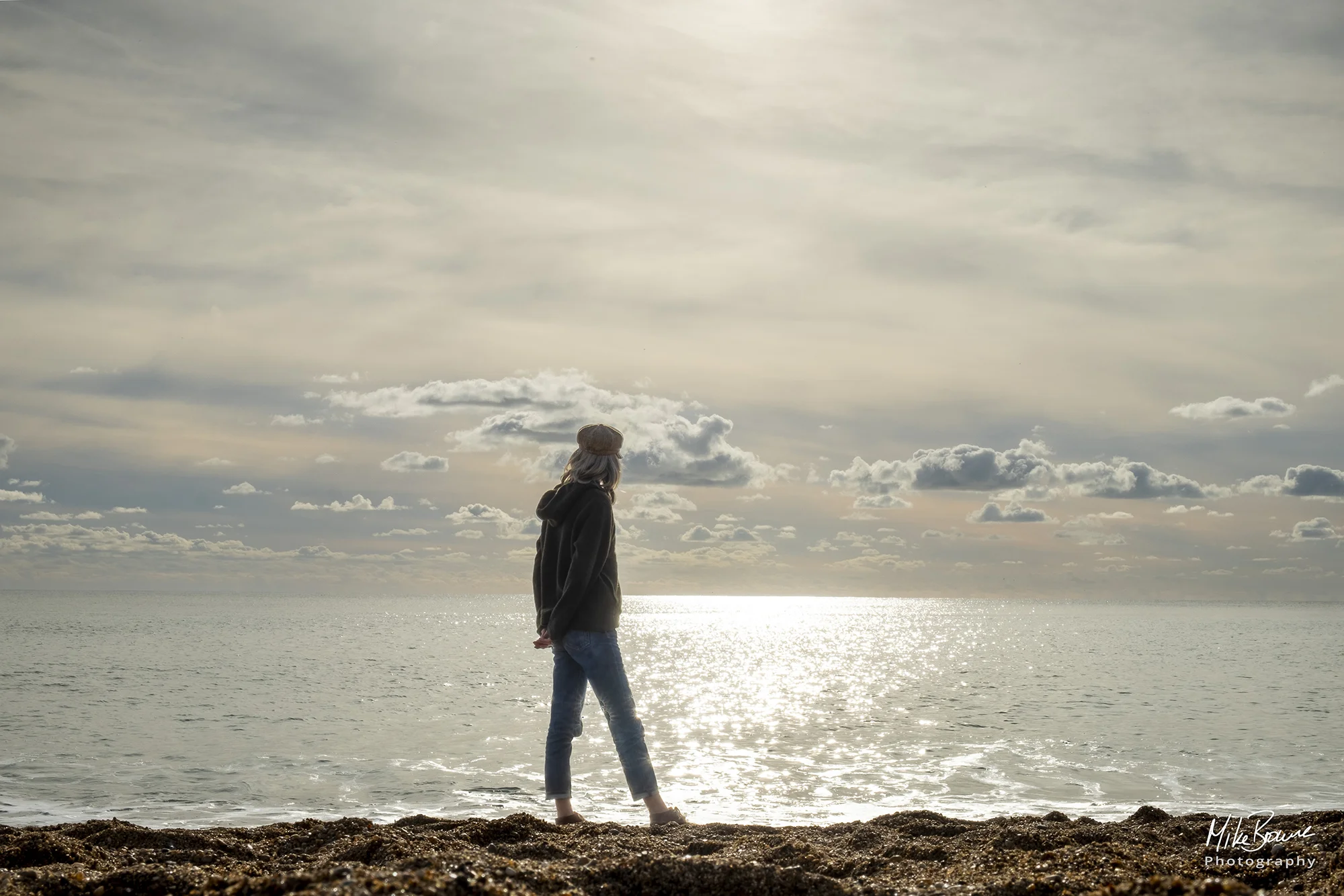 Slender woman wearing a wool cap with hands clasped behind her back looking at the sea and cloudy sky