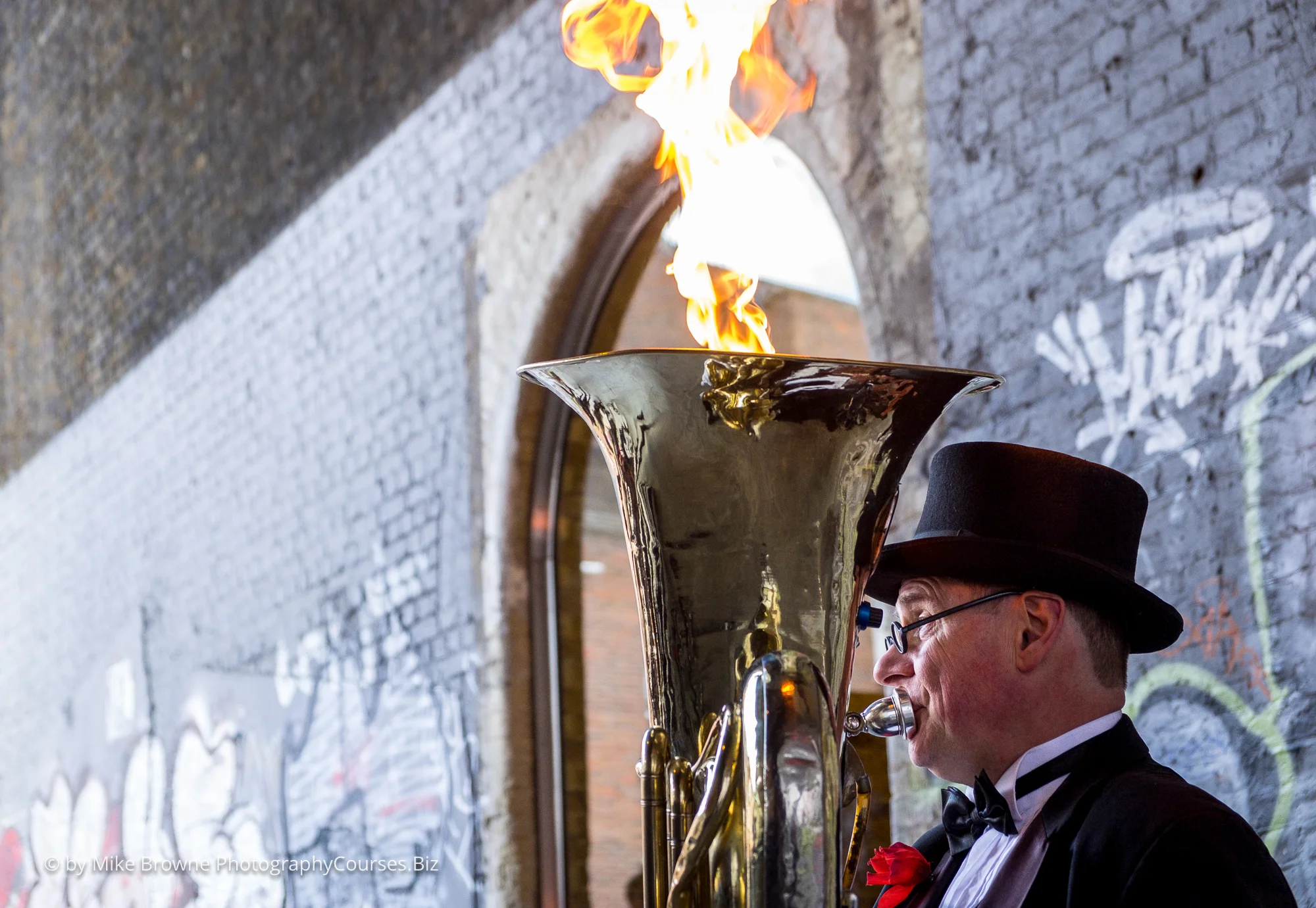 Man in top hat and bow tie playing the tuba in front of tagged brick wall