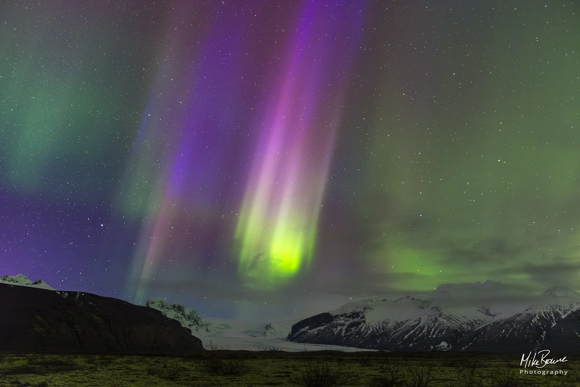 Streaks of magenta and green northern lights above snow and mountains