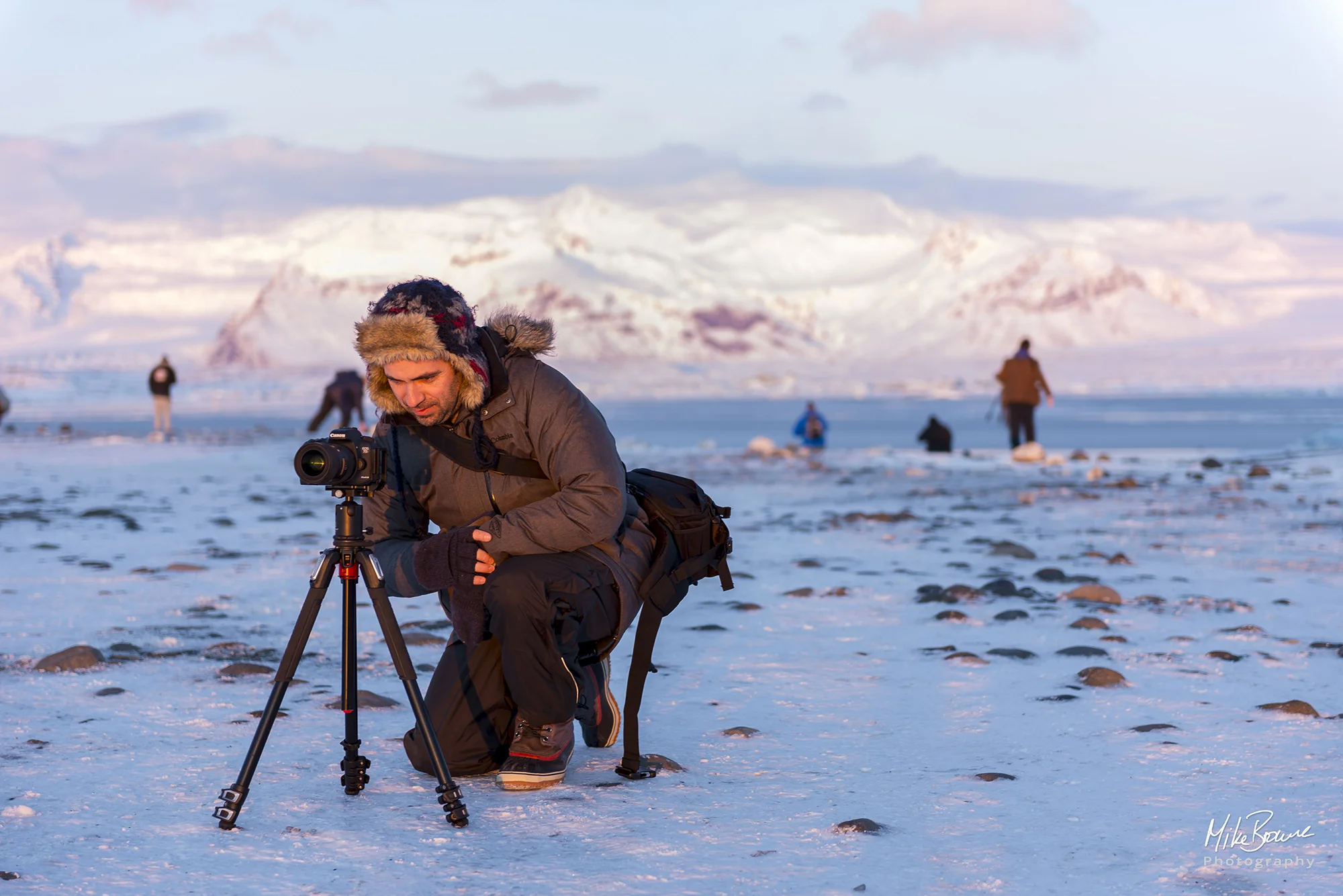Photographer crouching by camera and tripod with snow covered mountains behind