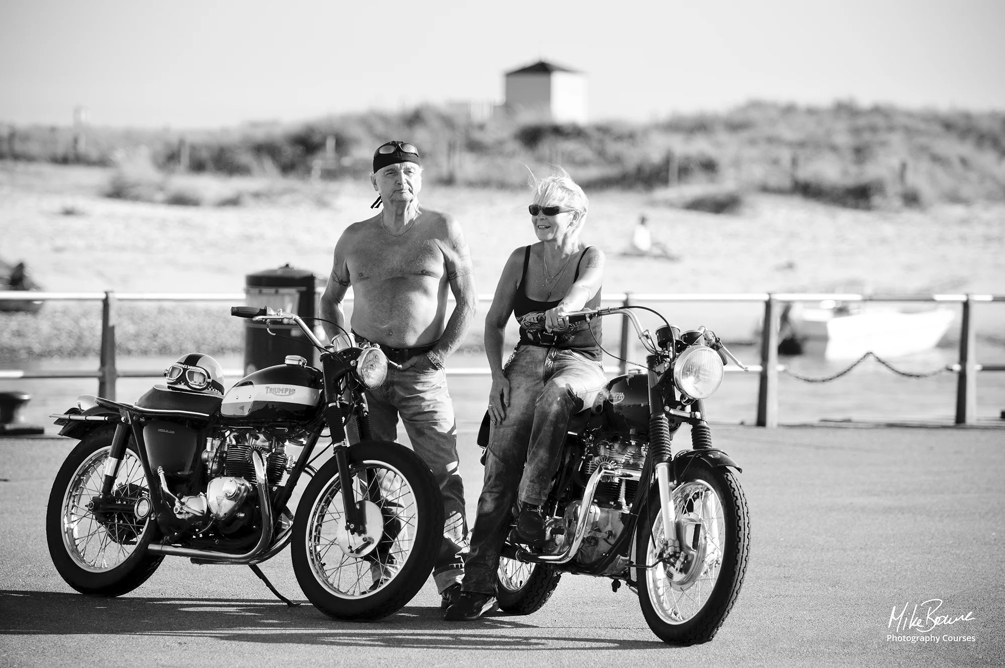 Older man and woman posing with vintage motorcycles by a beach