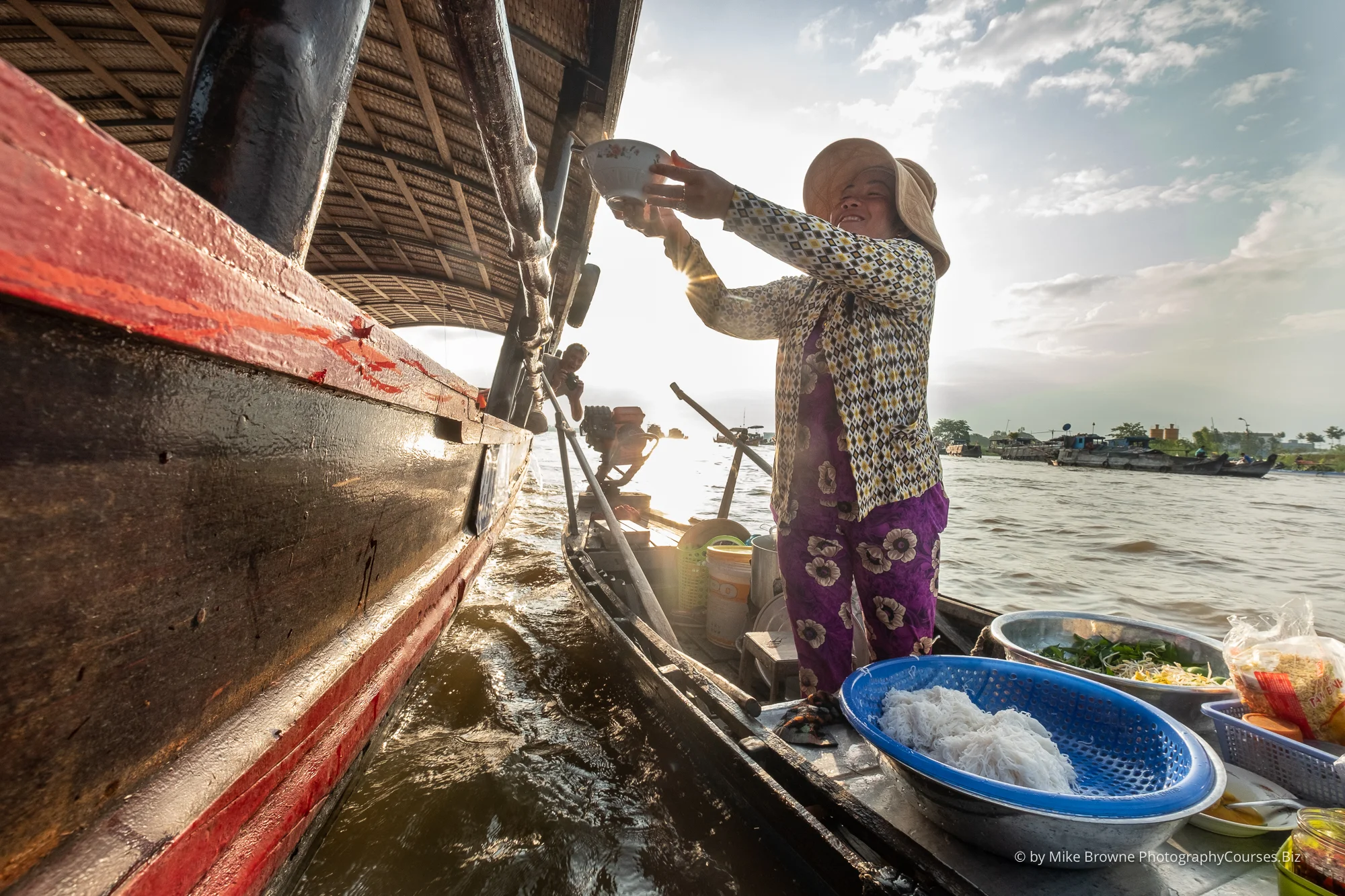 Woman standing in small boat passing food bowl up to another larger boat at Châu Đốc floating market, Vietnam