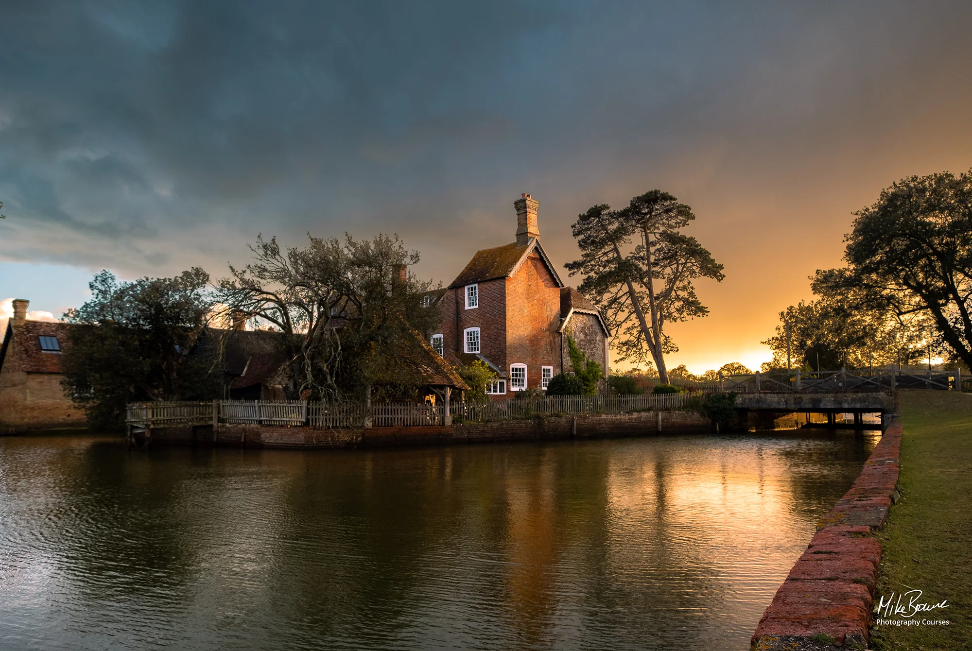 Tall red brick house and trees by a river at sunset