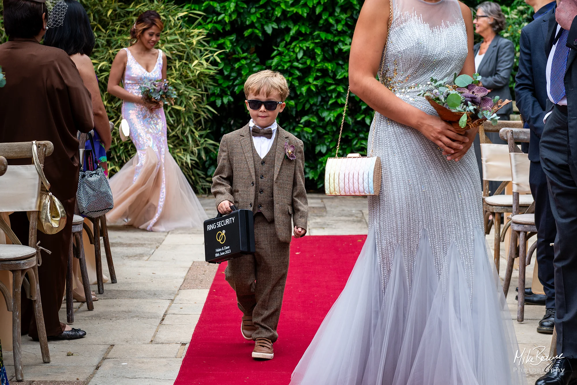 Page boy wearing a suit and sunglasses carrying attache case with Ring Bearer written on it