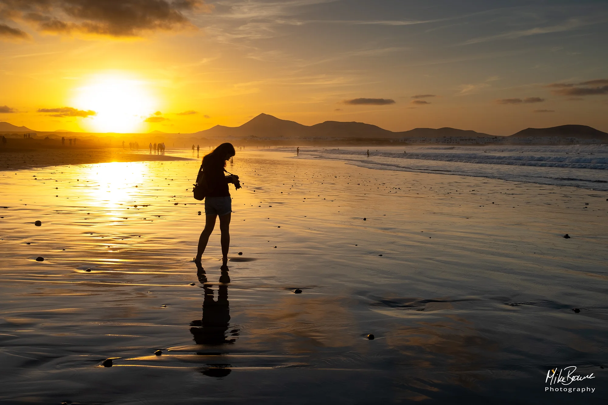 Female photographer standing on wet sand checking her camera at sunset, Famara Beach, Lanzarote