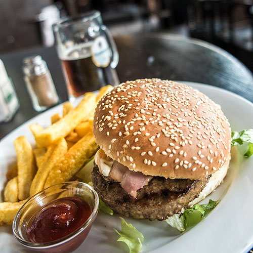 burger, chips and ketchup on a white plate with a few lettuce leaves scattered and a pint in the background