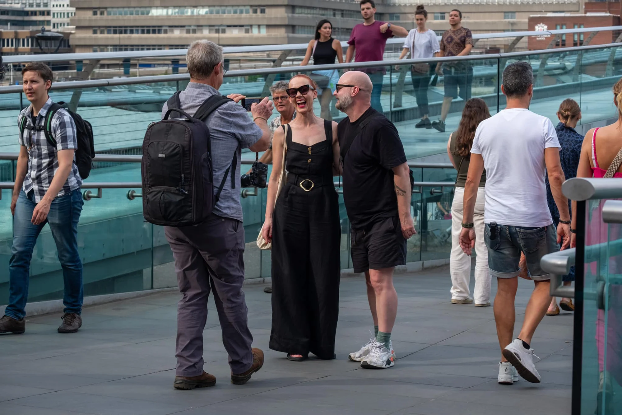 A photographer using a phone to take a photo of a man and woman wearing sunglasses on the Millennium Bridge in London