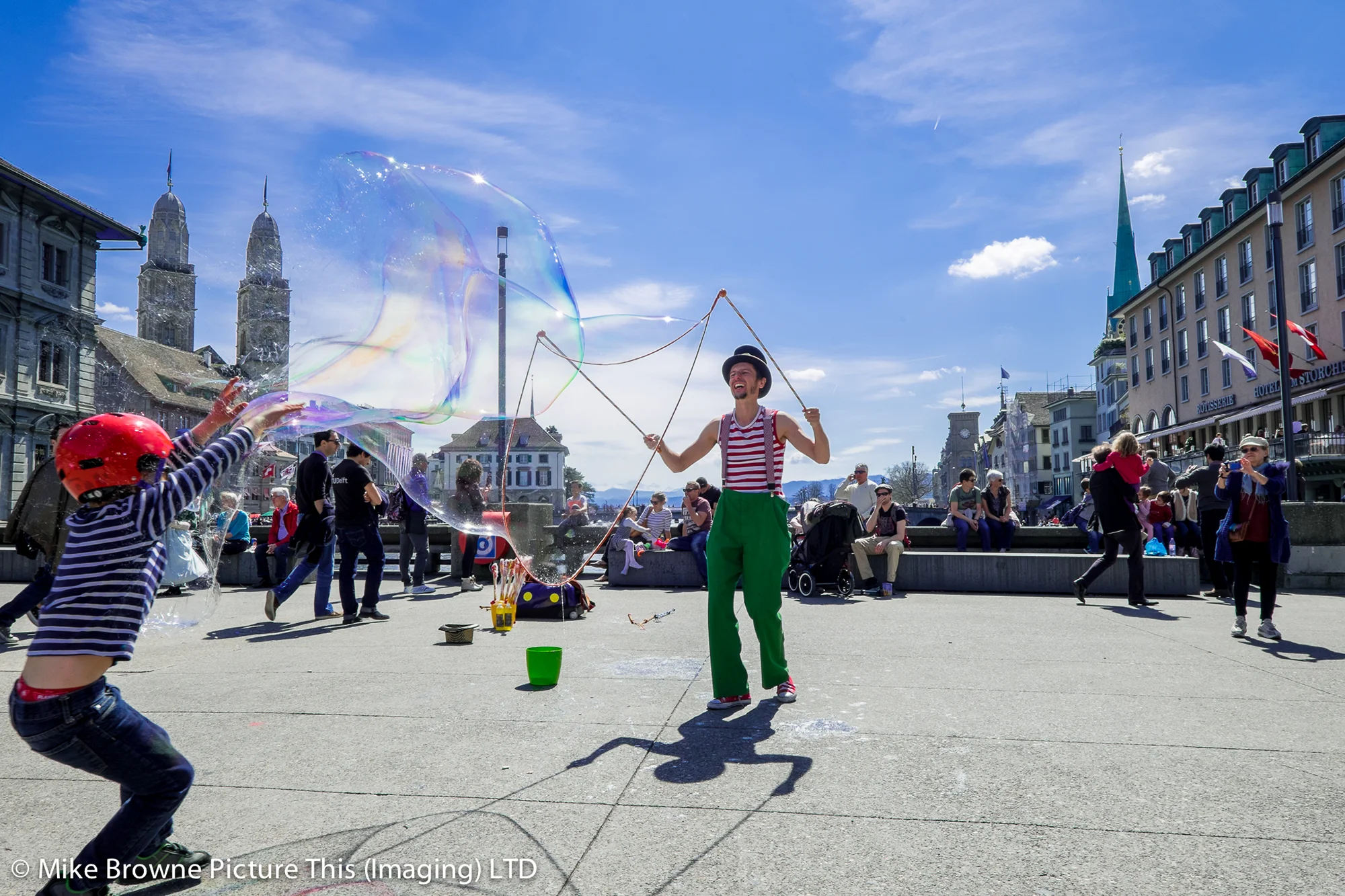 Man in clown costume on Rathausbrücke makes giant bubbles for children in the sunshine