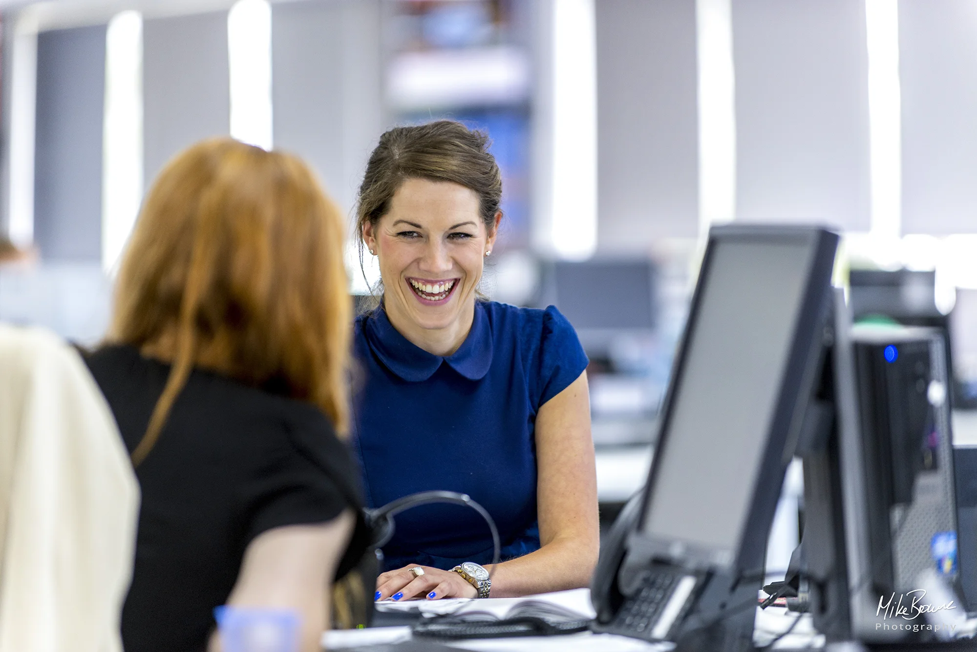 Woman wearing blue dress in an office environment in conversation to another woman with back to the camera
