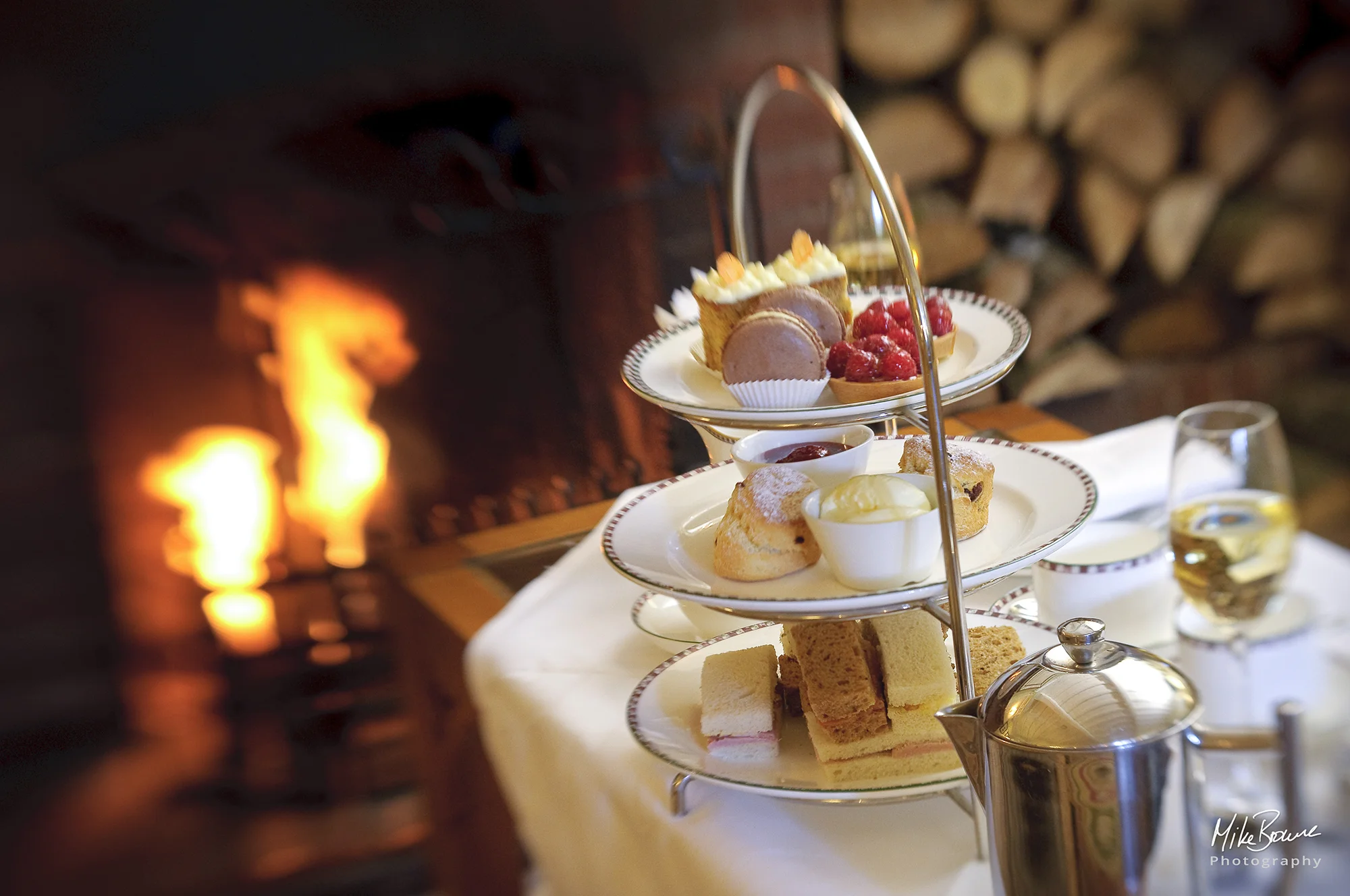 Classic English afternoon tea of sandwiches, cakes, scones and jam with cream and pot of tea next to log fire