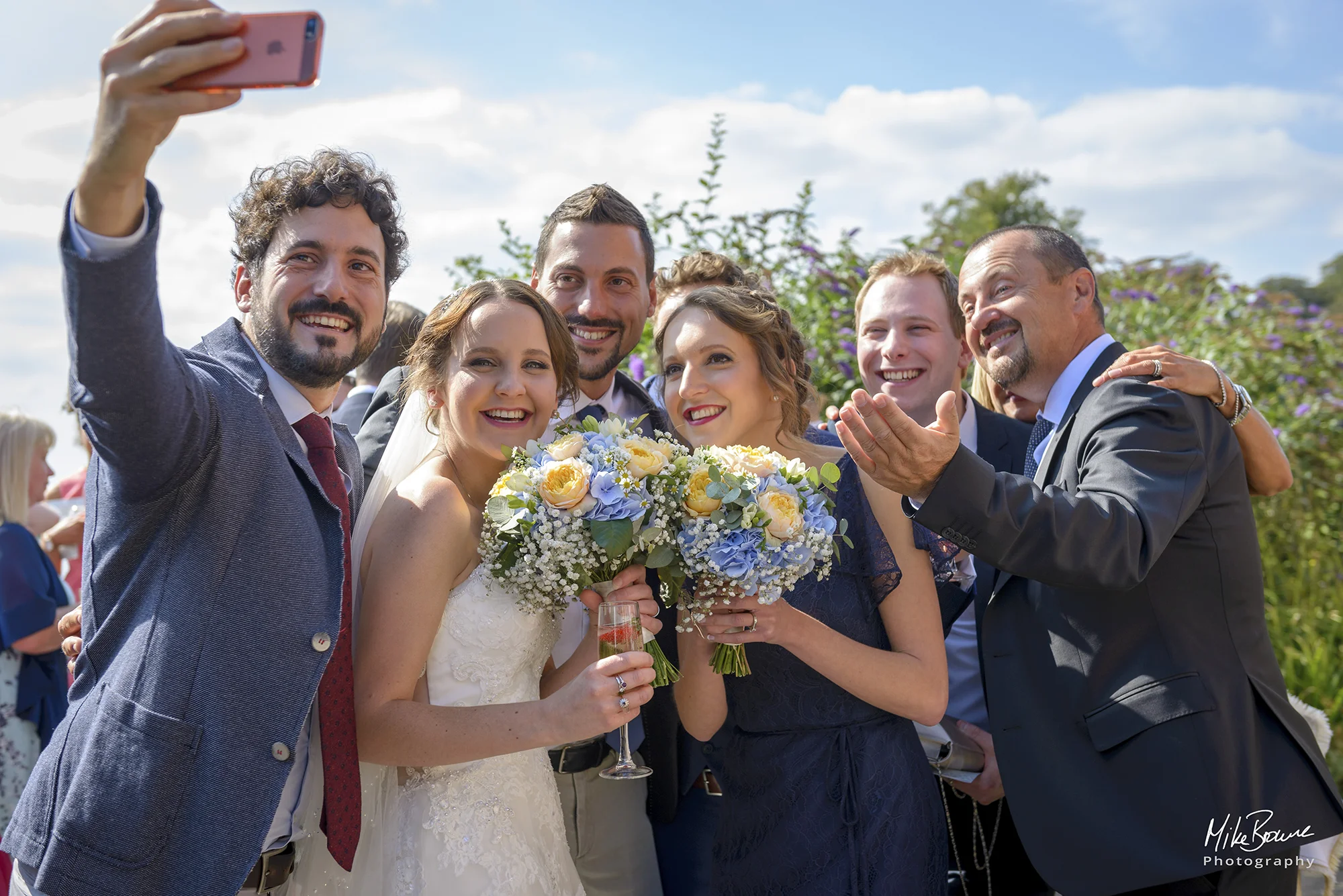 Bride and bridesmaid holding bouquets posing for a selfie with friends on a sunny day