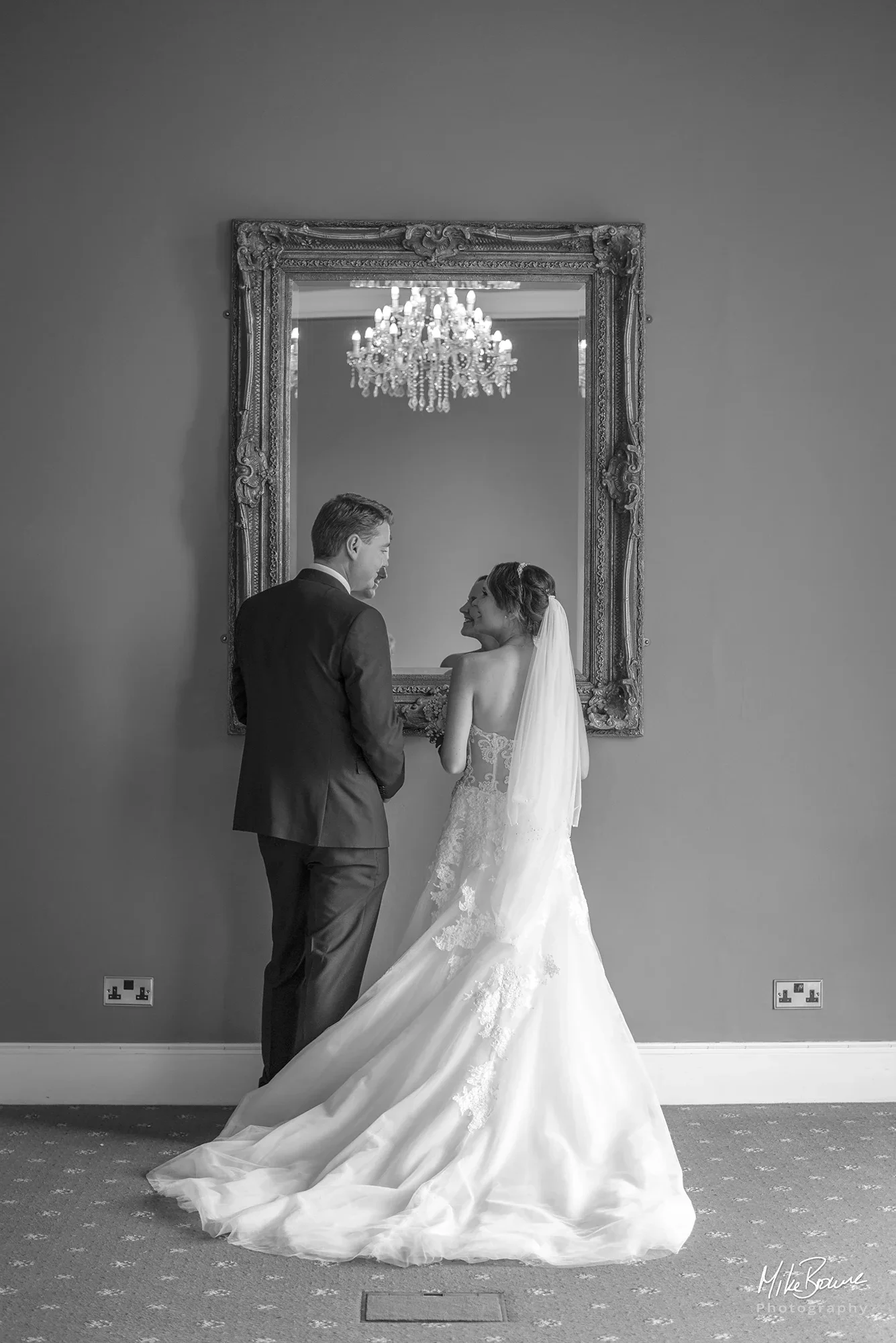 Newly wedded couple looking at each other in front of a large wall mirror with reflection of a chandelier