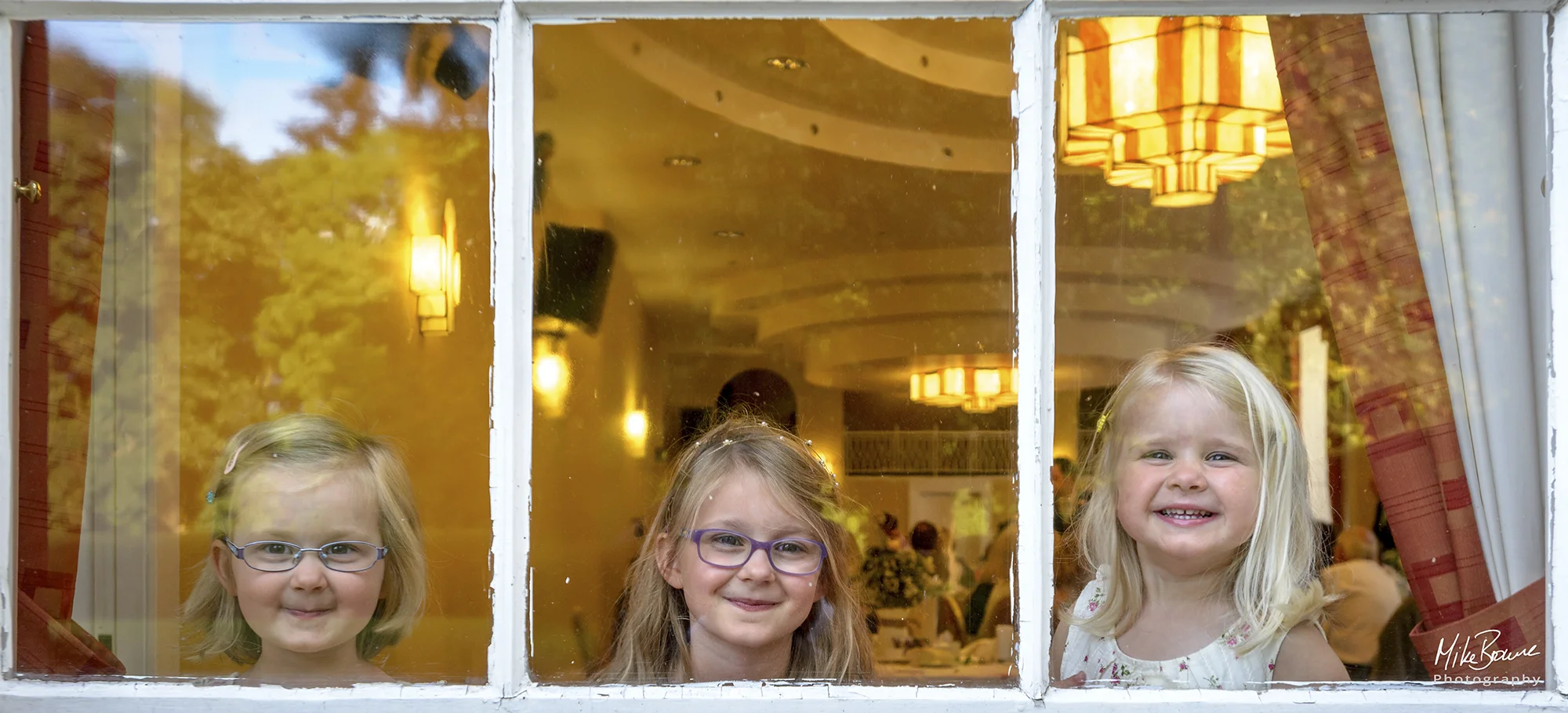 three young flower girls looking out of a window at a wedding reception