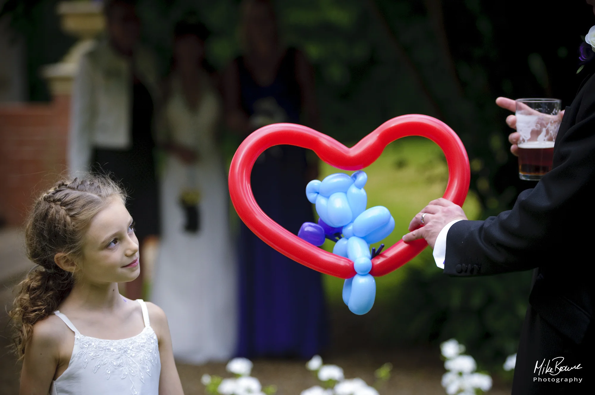 A young bridesmaid gazes quizzically at a balloon model of a blue teddy bear sitting in a red heart