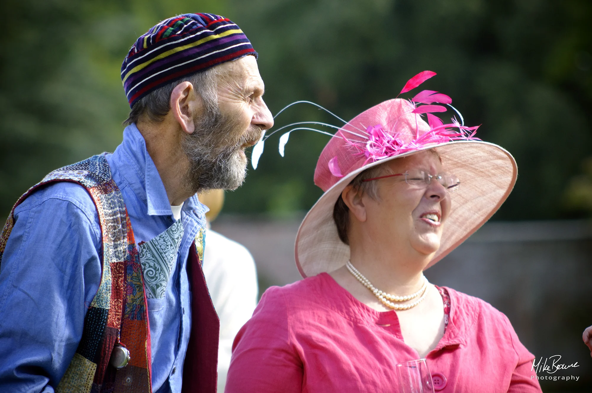 Bearded man wearing new age clothes laughing with a woman in a pink hat with feathers