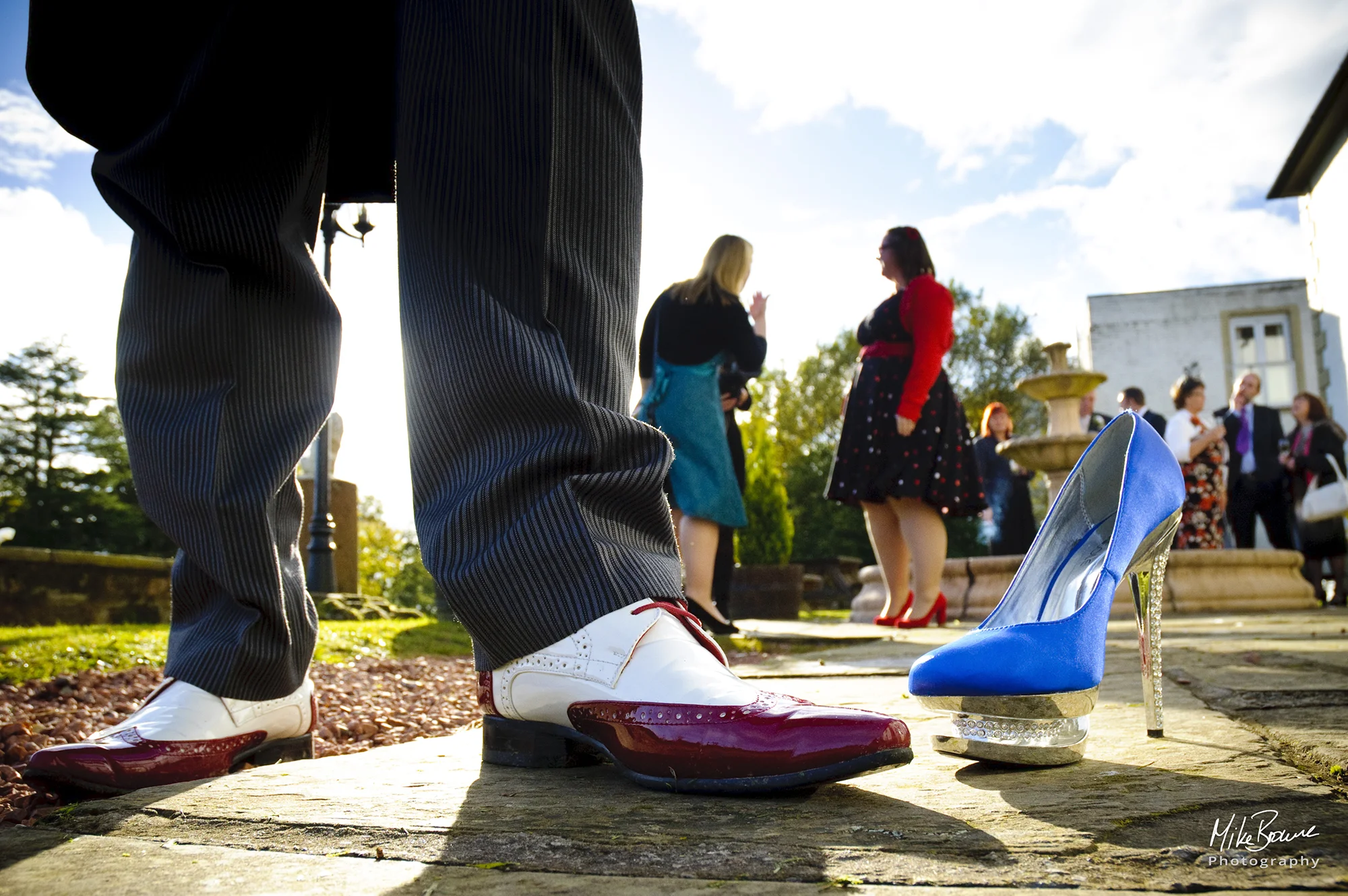 Man\'s pinstripe trouser legs with trendy white and maroon brogue shoes by a blue and chrome stiletto shoe and people milling about in the background at a reception