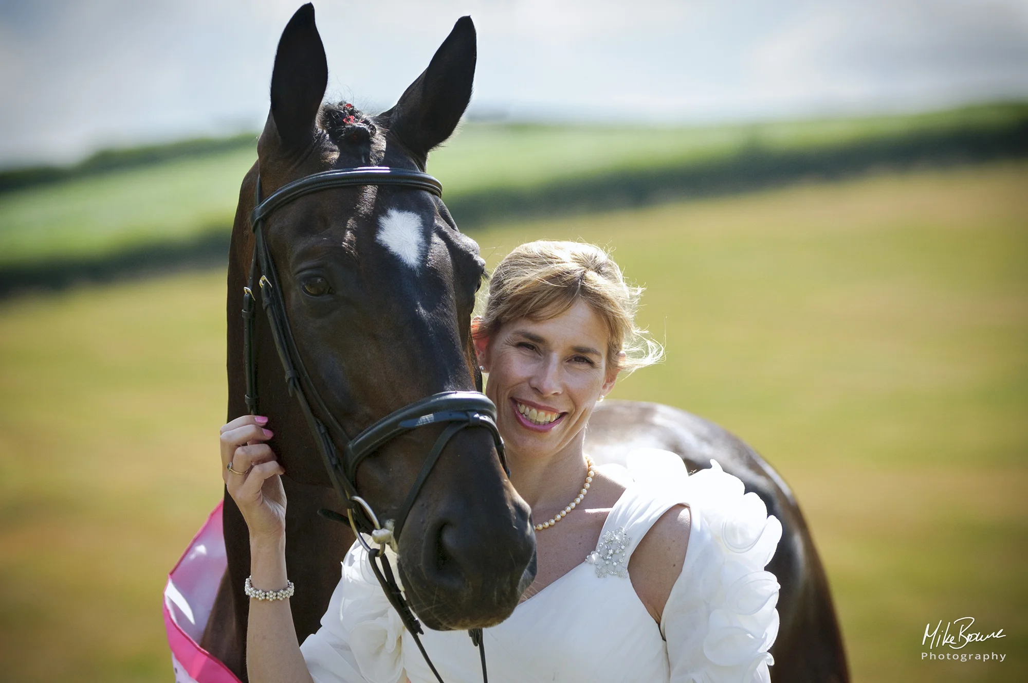 Smiling woman in a wedding dress holding the head of a thoroughbred horse in a field on a sunny day