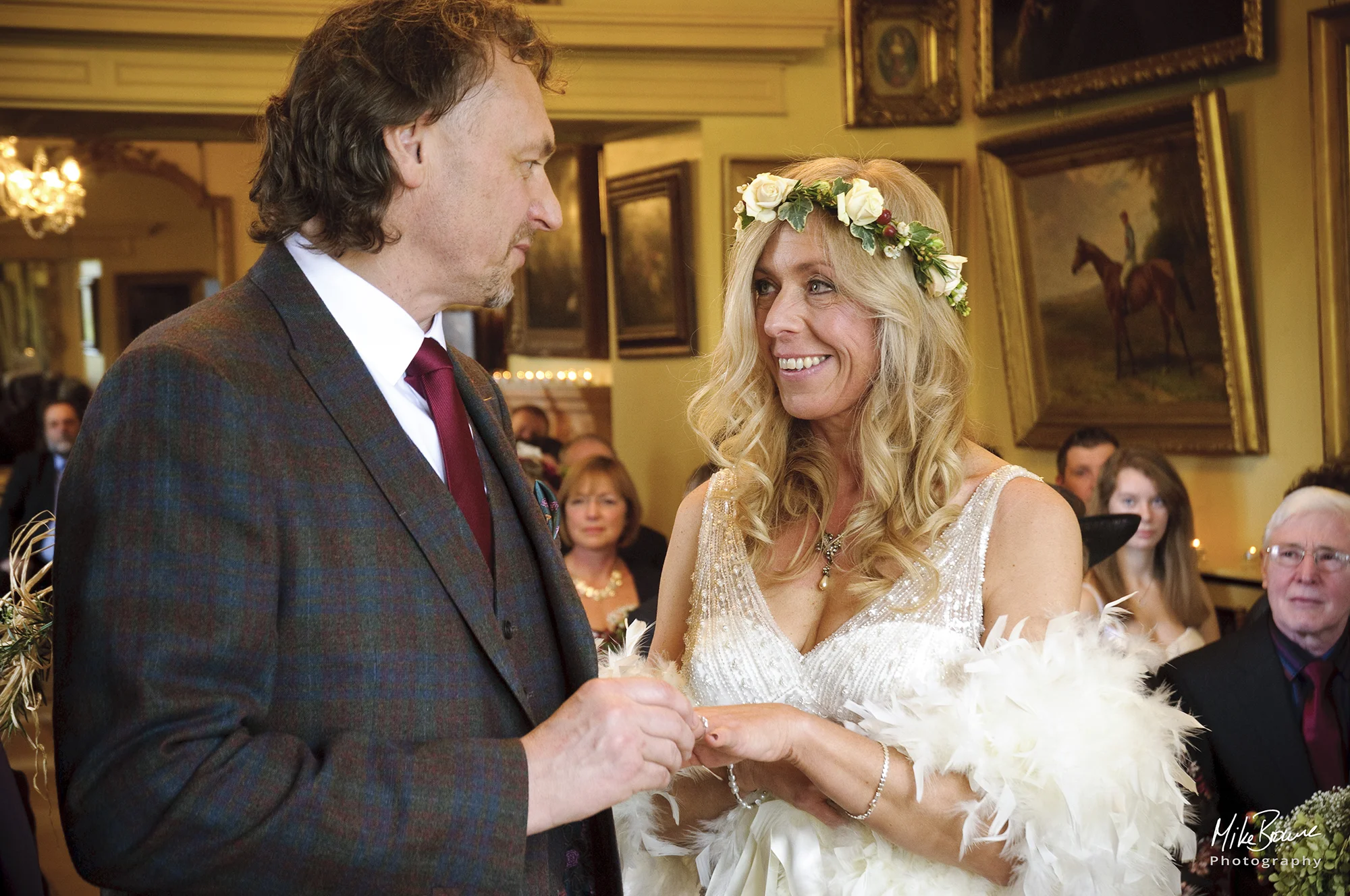 Man in a brown check suit and a woman wearing a wedding dress and flowers in her hair look at each other as he places a ring on her finger