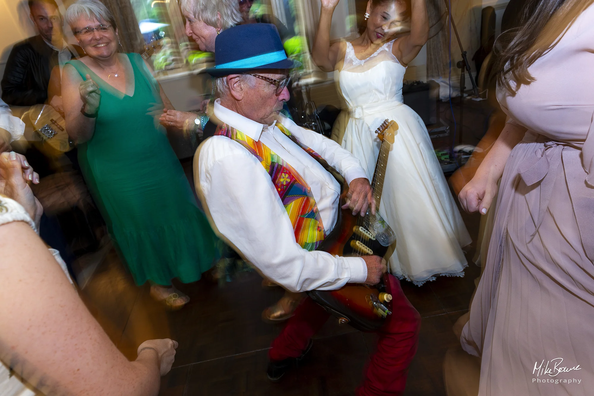 Man in colourful clothes plays guitar to a bride as people dance around them
