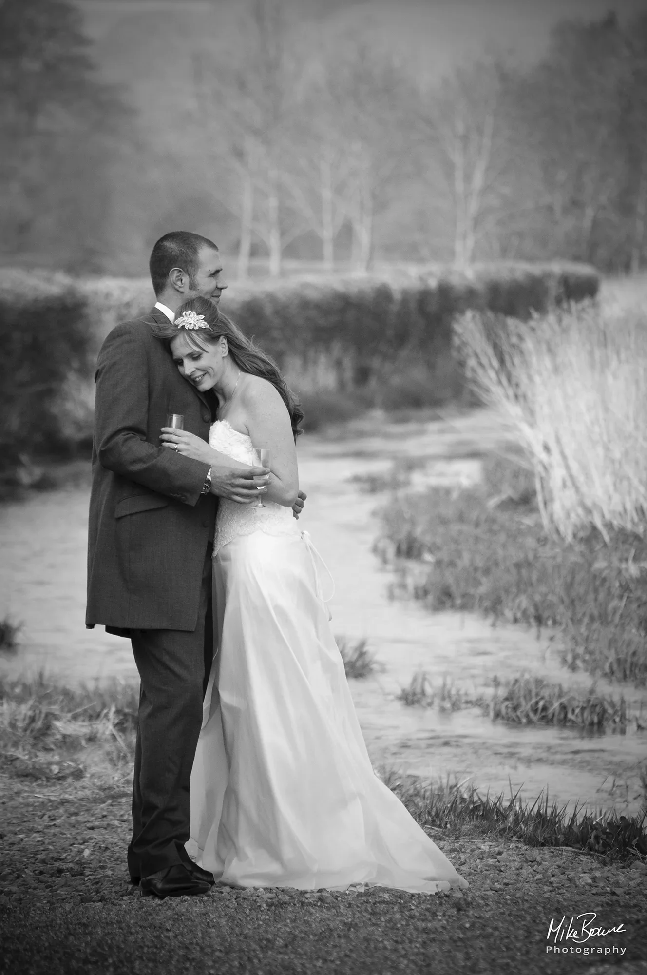 Man in a morning suit and woman in a wedding dress with glasses of Champagne embracing next to a river