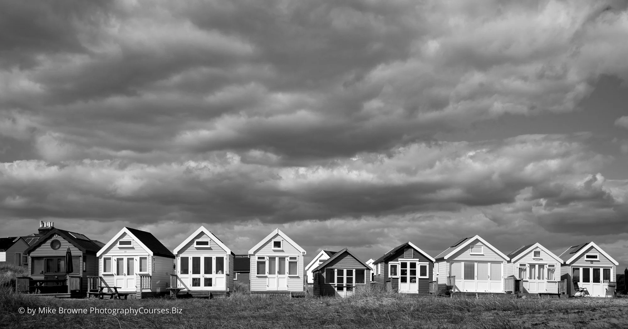 Row of nine beach huts in sunshine with storm clouds in sky