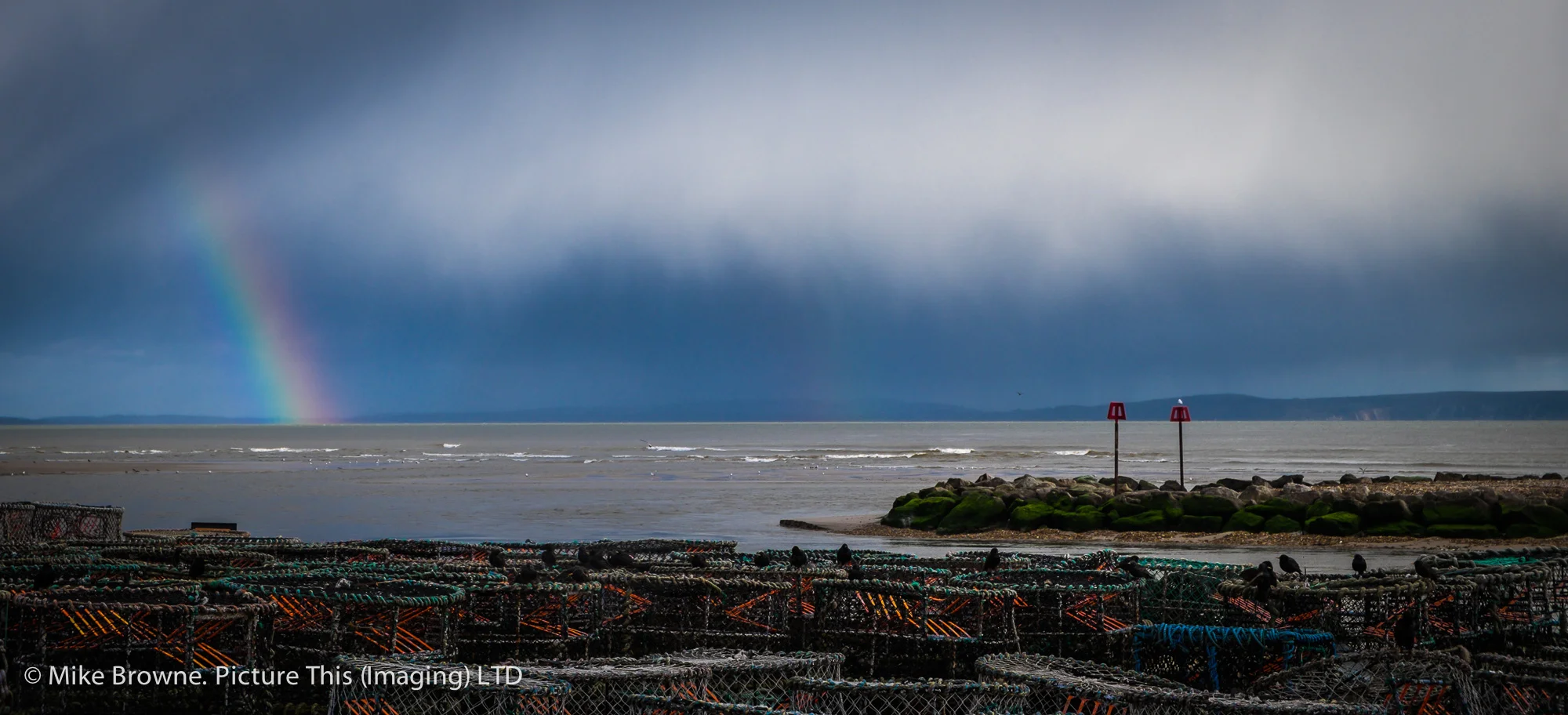 Lobster pots on a quayside and a rainbow over the sea behind them