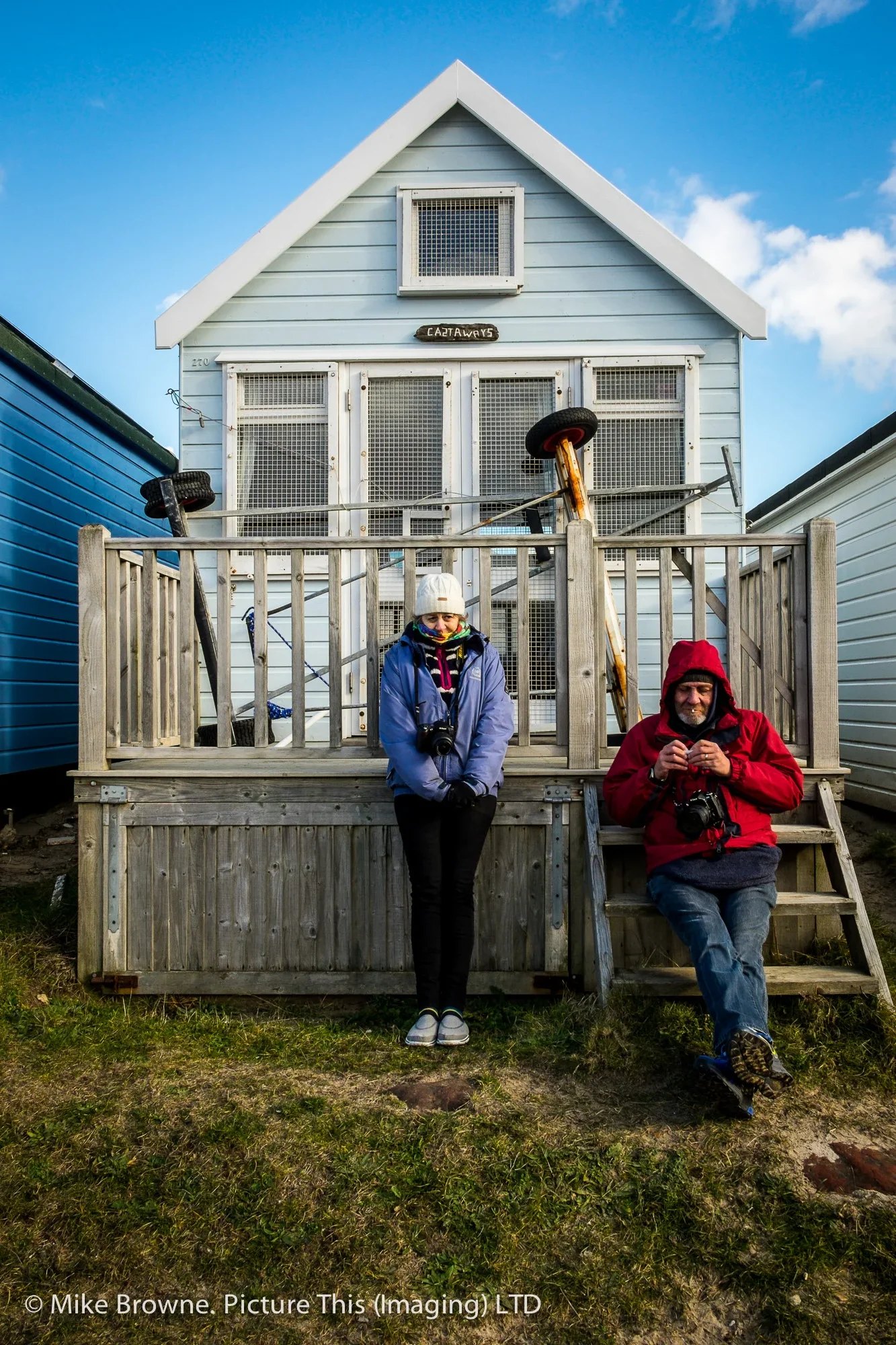 Man and woman dressed in winter clothing looking at cameras and resting against a blue British beach hut