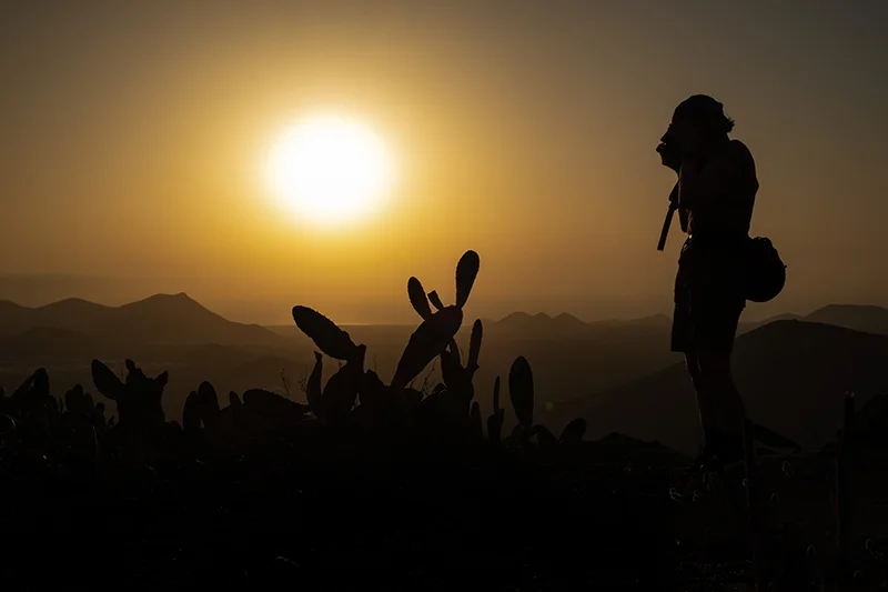 Female photographer photographing a cactus silhouetted against hazy orange sunset with distant mountains